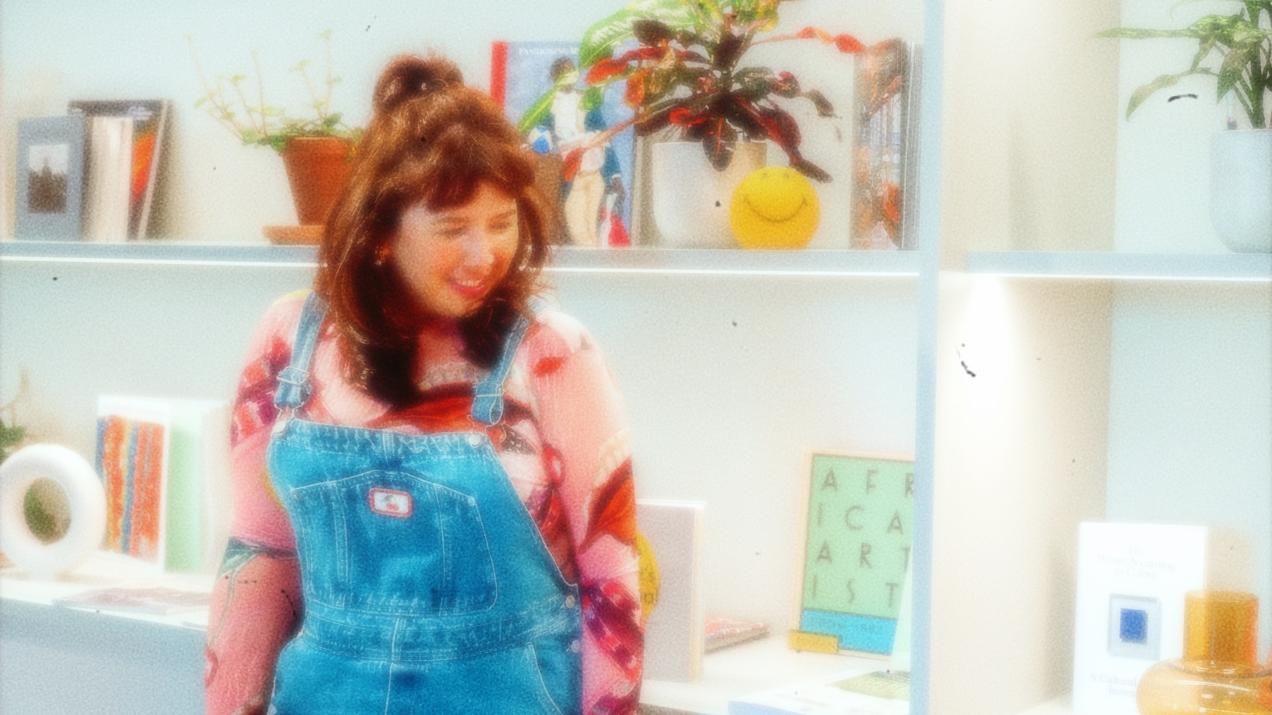 A woman with red hair wearing a pink shirt and blue overalls, smiling, standing in front of a bookshelf decorated with plants and books.