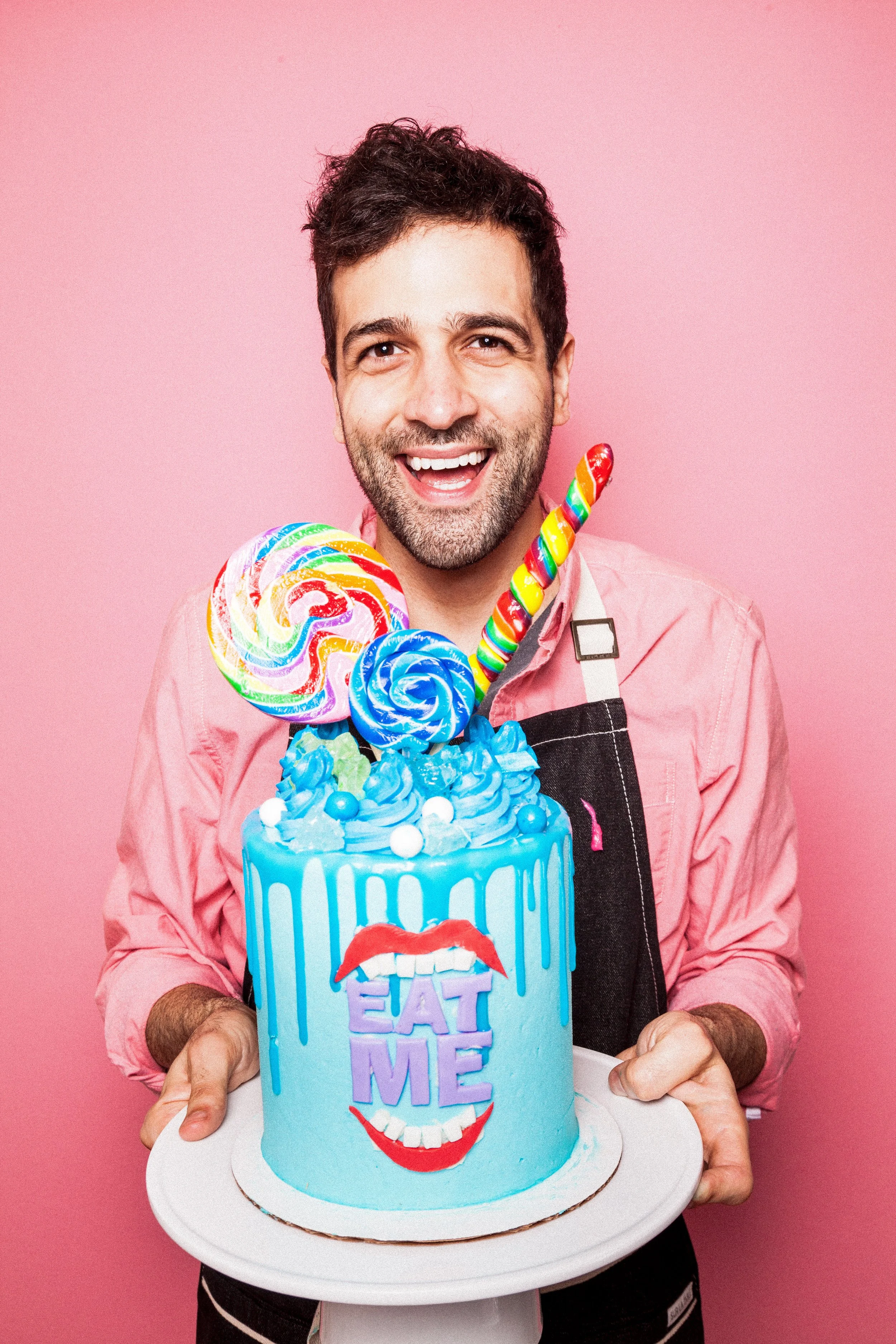 A man holding a birthday cake decorated with blue icing, colorful lollipops, and a face with red lips and a tongue out, against a pink background.