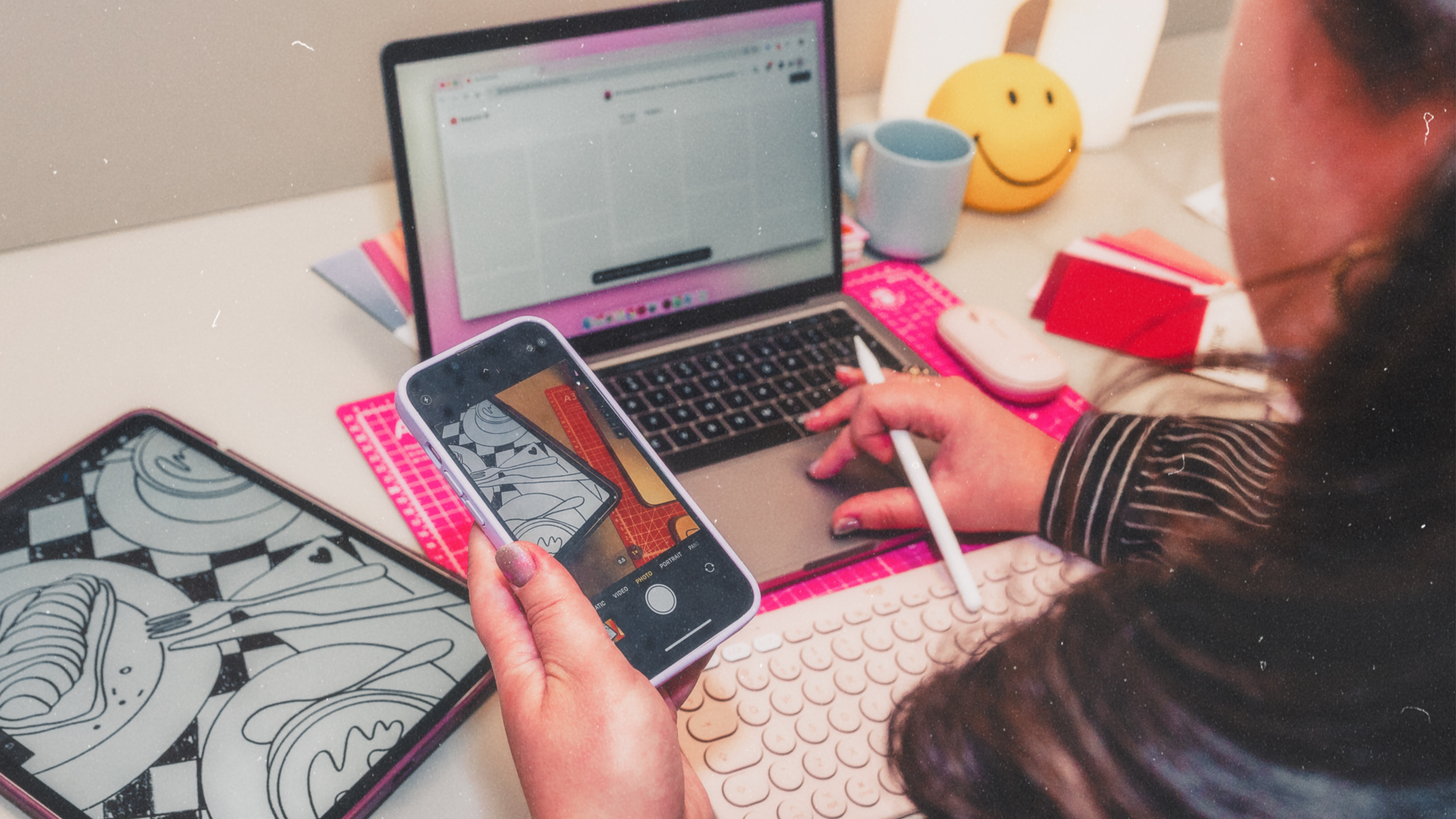A person holding a smartphone and taking a picture of a workspace with a laptop, sketchpad, mug, and a smiling yellow emoji lamp.