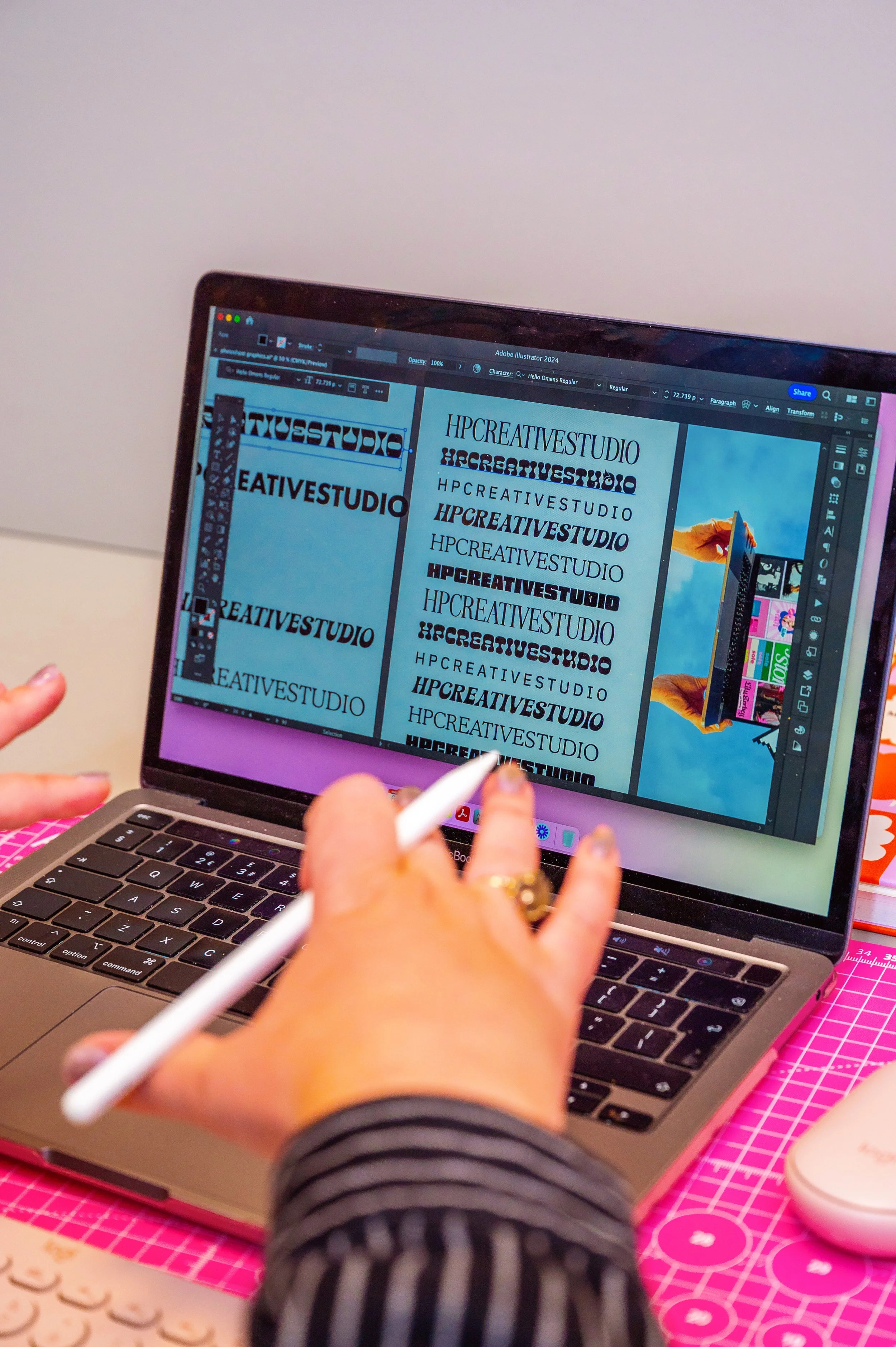A person working on a laptop with a design software open, displaying various text fonts for 'HPCREATIVESTUDIO' on the screen. The workspace includes a pink cutting mat with a grid pattern underneath the laptop and some colorful items nearby.