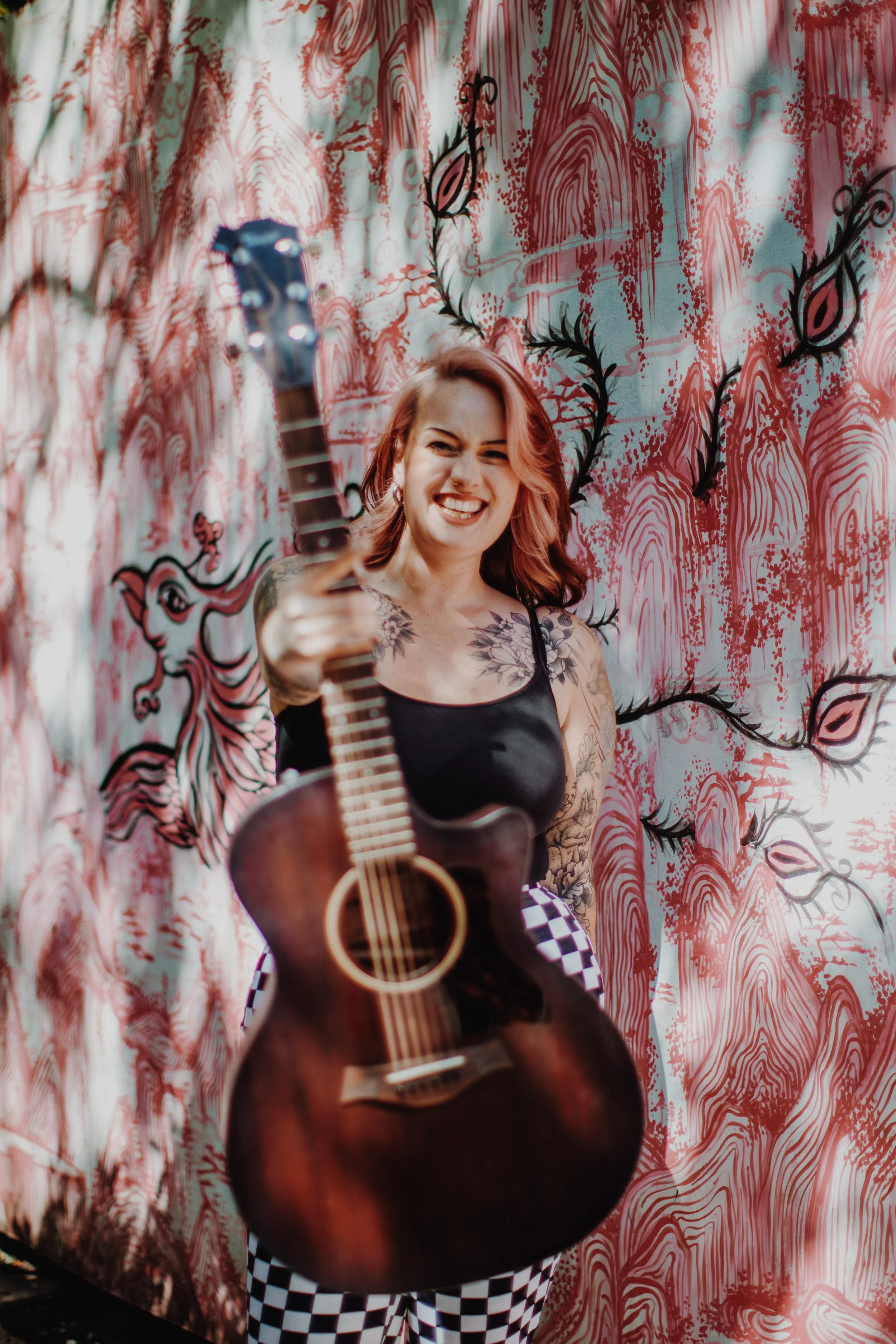 Woman with wavy pink hair holding an acoustic guitar and smiling in front of a colorful, patterned wall.