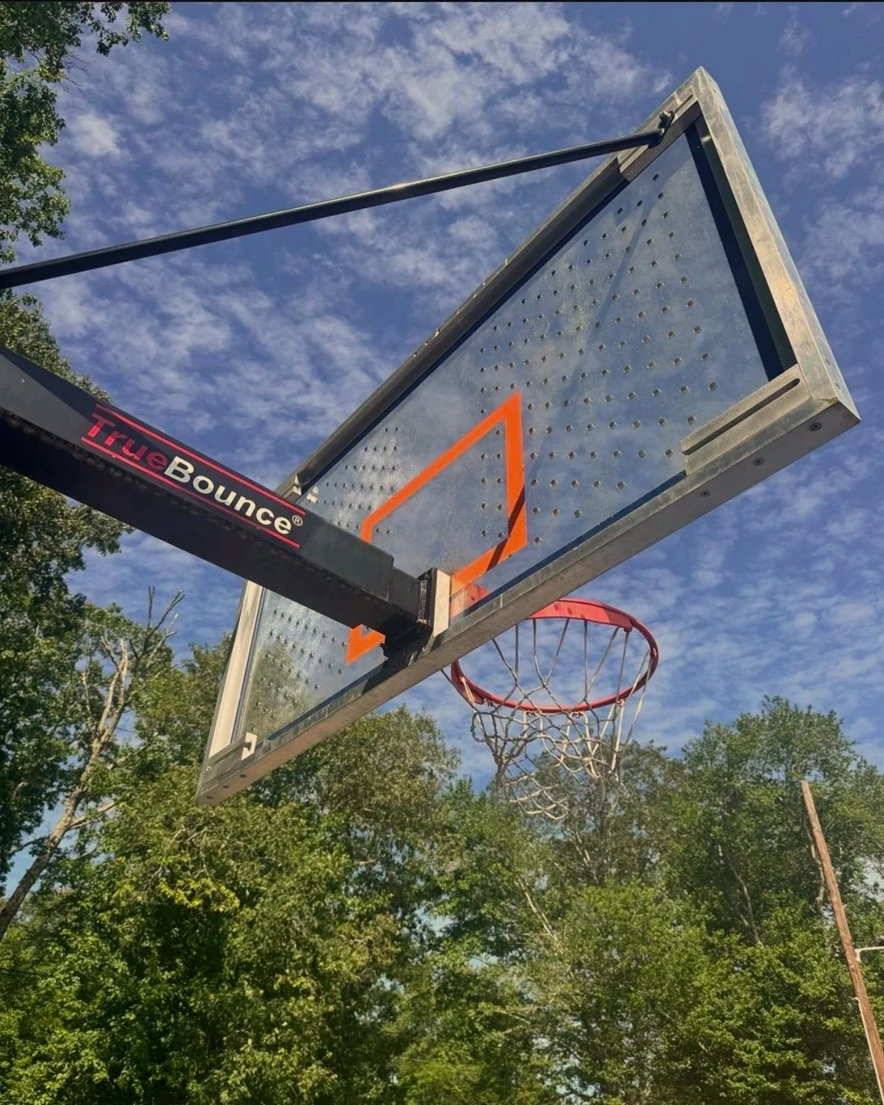 Outdoor basketball hoop with backboard and net, shot from below against a partly cloudy sky and green trees in the background.