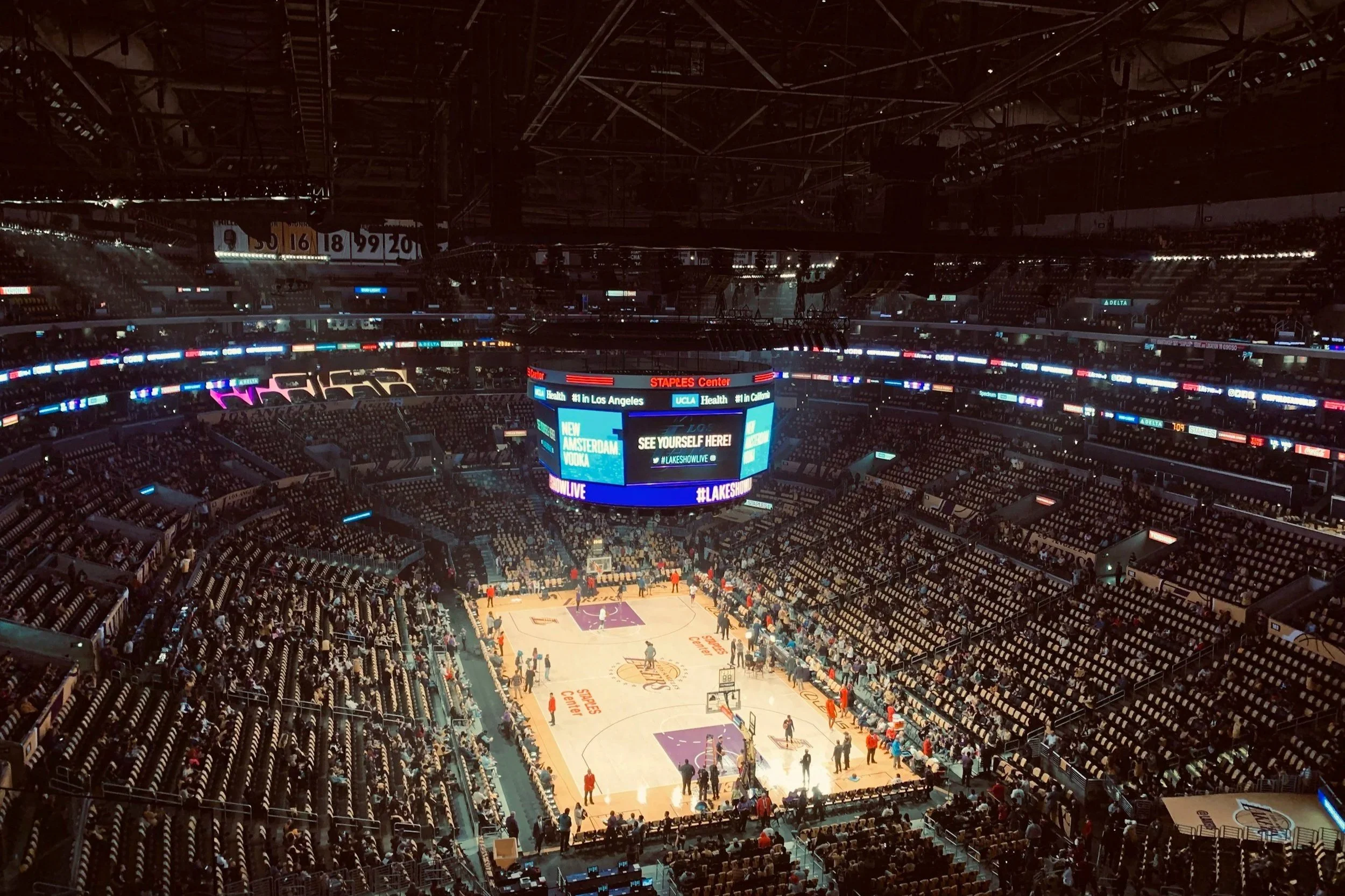 Inside an empty basketball arena with a few players warming up on the court, surrounded by rows of seats, large electronic scoreboard overhead displaying advertisements and messages.