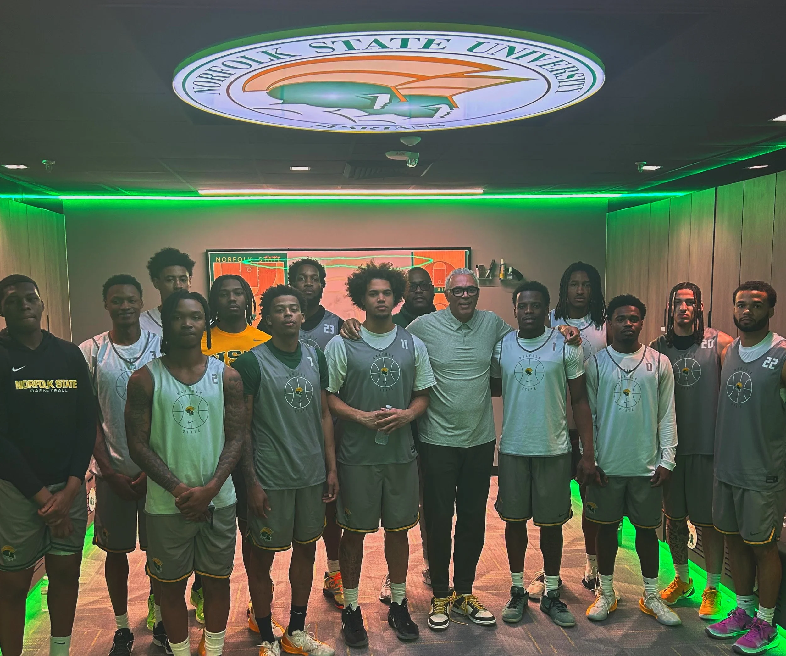 Group of young basketball players and coach posing for a team photo at Norfolk State University basketball court with signage overhead.
