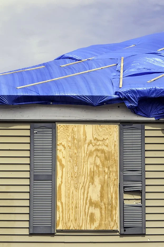 A house with a boarded-up window and shutters, and a blue tarp covering the roof, services by The Board Up Company in St. Louis MO