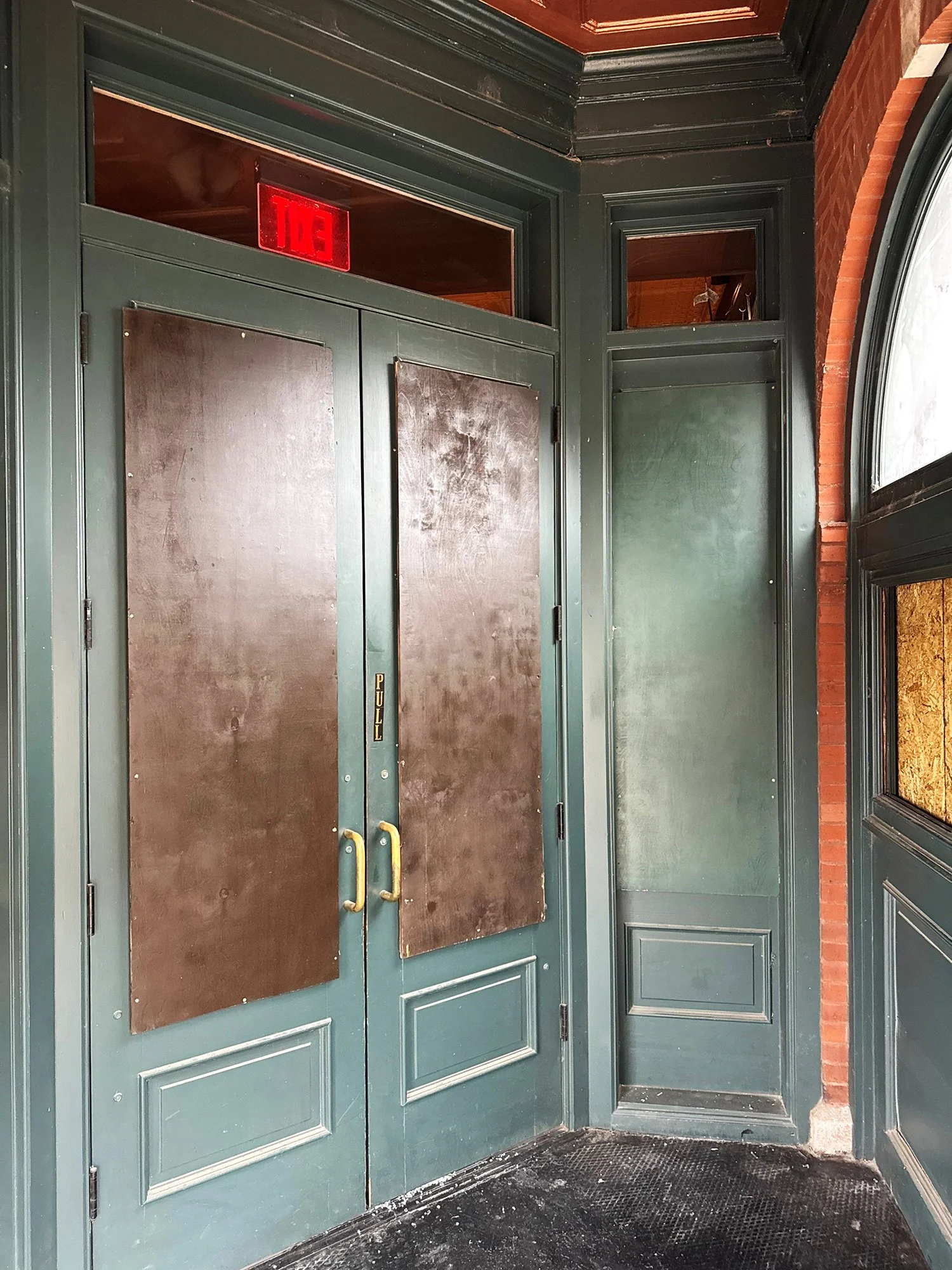 Interior of an entrance with dark green double doors with brass handles, a pull sign, and a red-lit exit sign above. The doors have wood from boarding up services by The Board Up Company in St. Louis MO