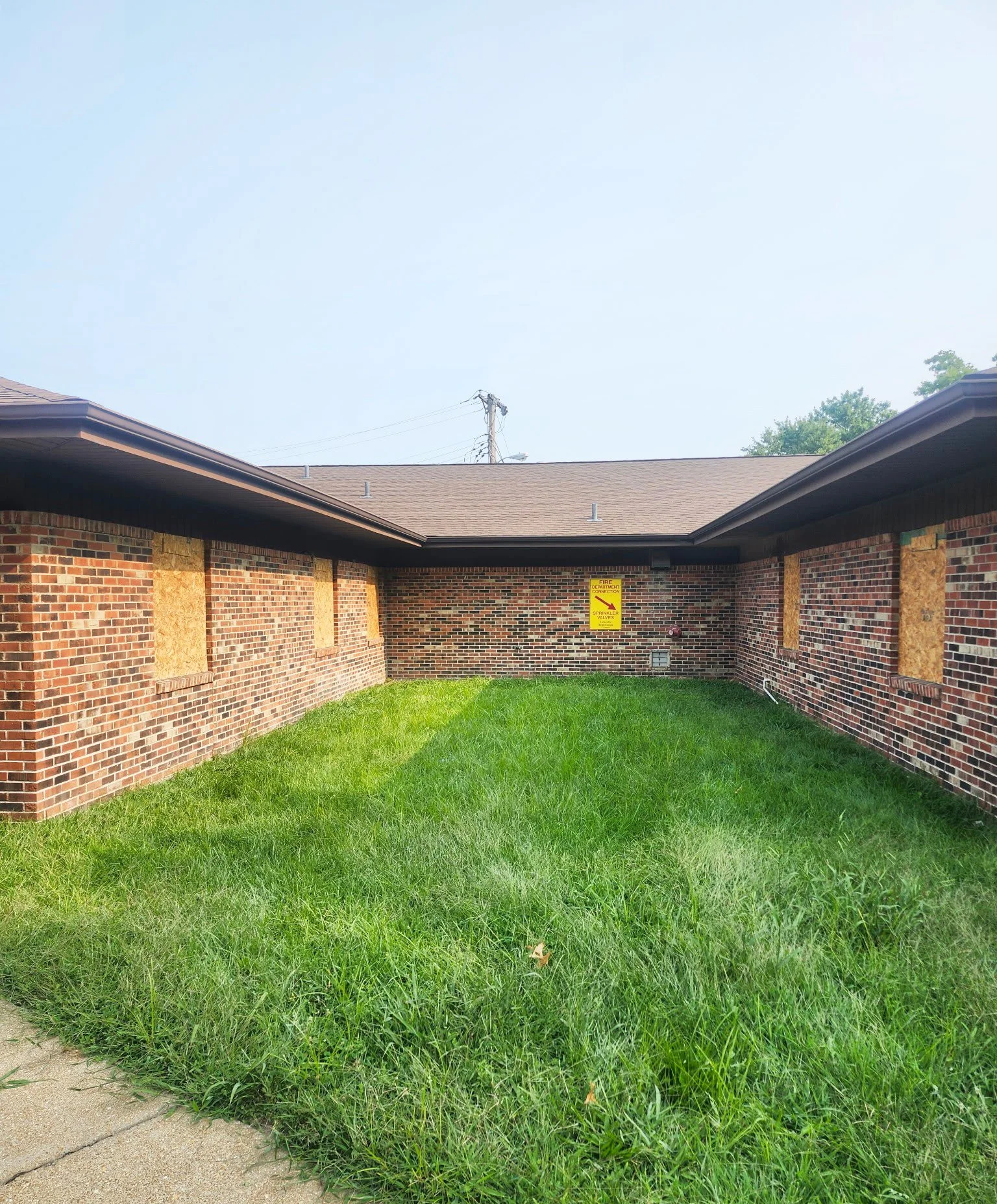 Backyard of a brick building with boarded-up windows by The Board Up Company in St Louis Missouri, a green grassy area, and a sidewalk in the foreground, under a clear blue sky.
