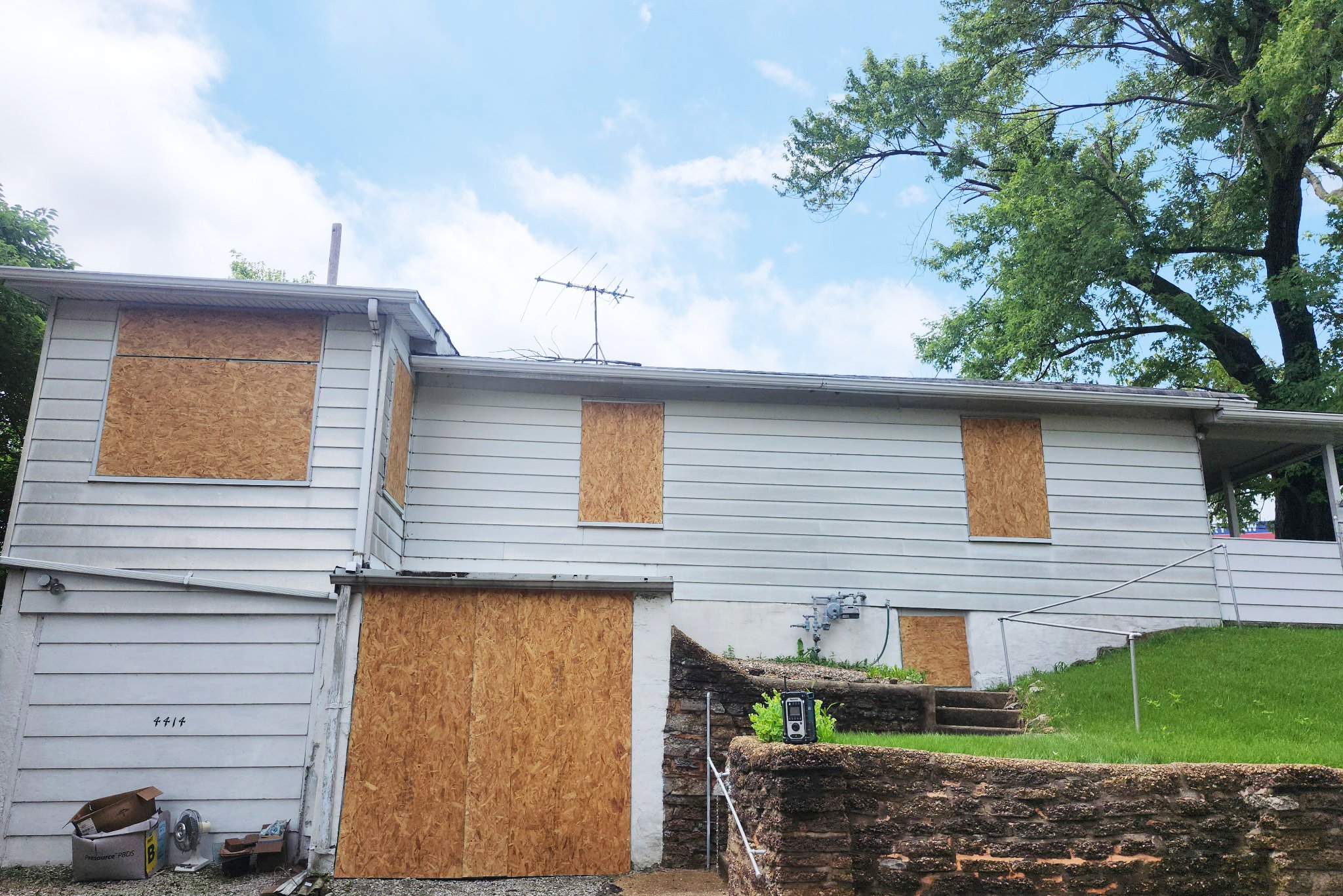 A two-story house with boarded-up windows on the upper and lower levels, white siding, and a satellite dish on the roof. Residential property after damage to windows and doors with boarding up services by The Board Up Company in St. Louis MO.