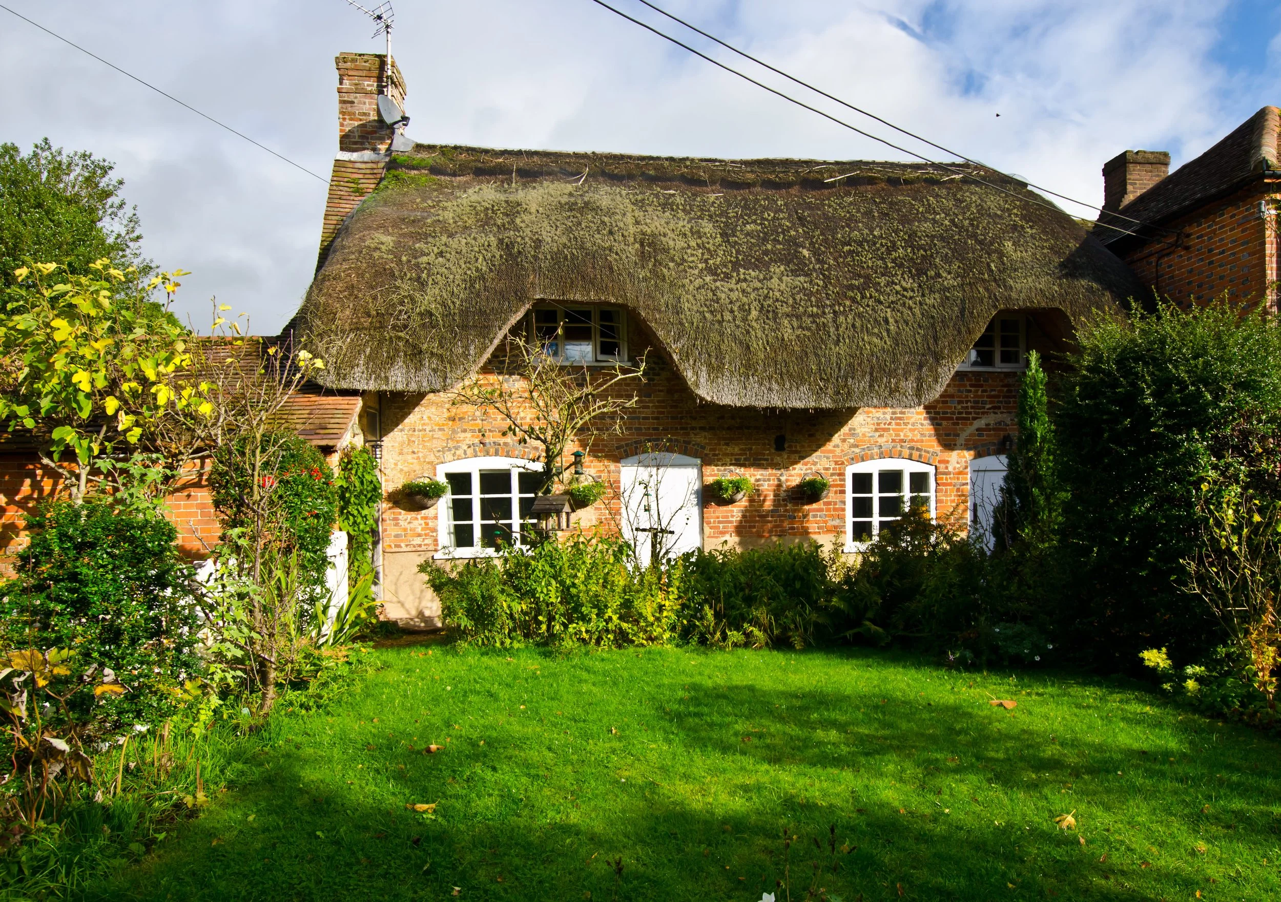 A thatched-roof brick house surrounded by a well-maintained garden with green grass, flowering plants, and trees, under a partly cloudy sky.