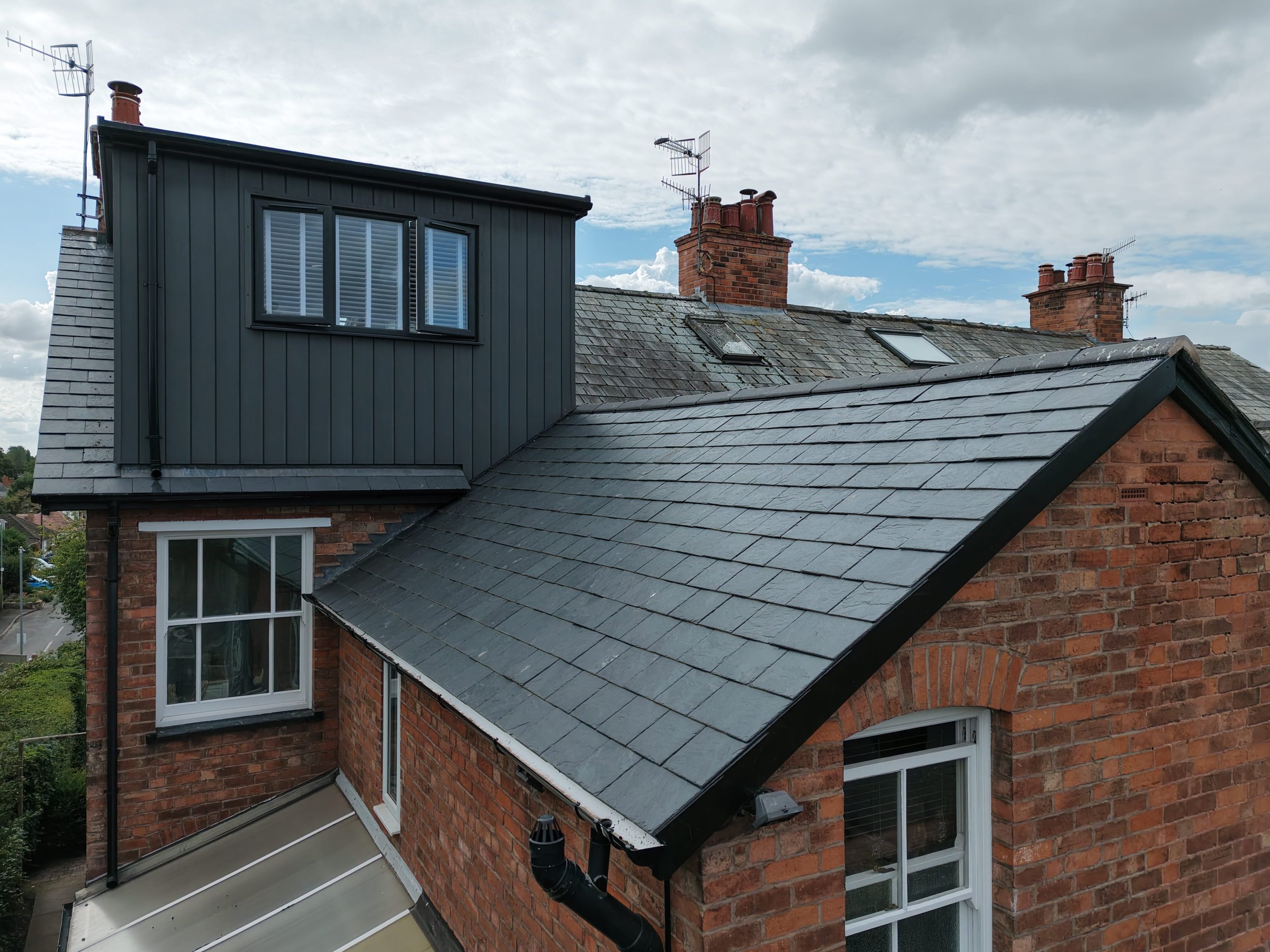The rooftops of a red brick house with multiple chimneys, skylights, and television antennas against a cloudy sky.