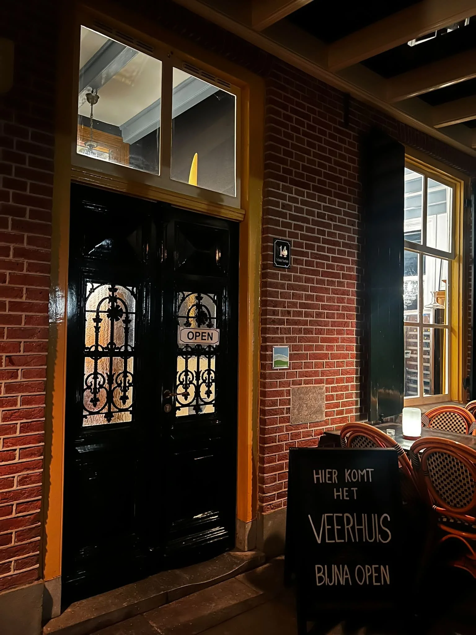 Een restaurant of café met een rode bakstenen muur, grote ramen met gele raampartijen, en een zwart metalen deur met een 'Open' bord. Binnen is het licht en er staan stoelen en tafels met borden en glazen. Een bord in de voorgrond zegt 'Hier komt het lekkerder veerhuis, bijna open'.