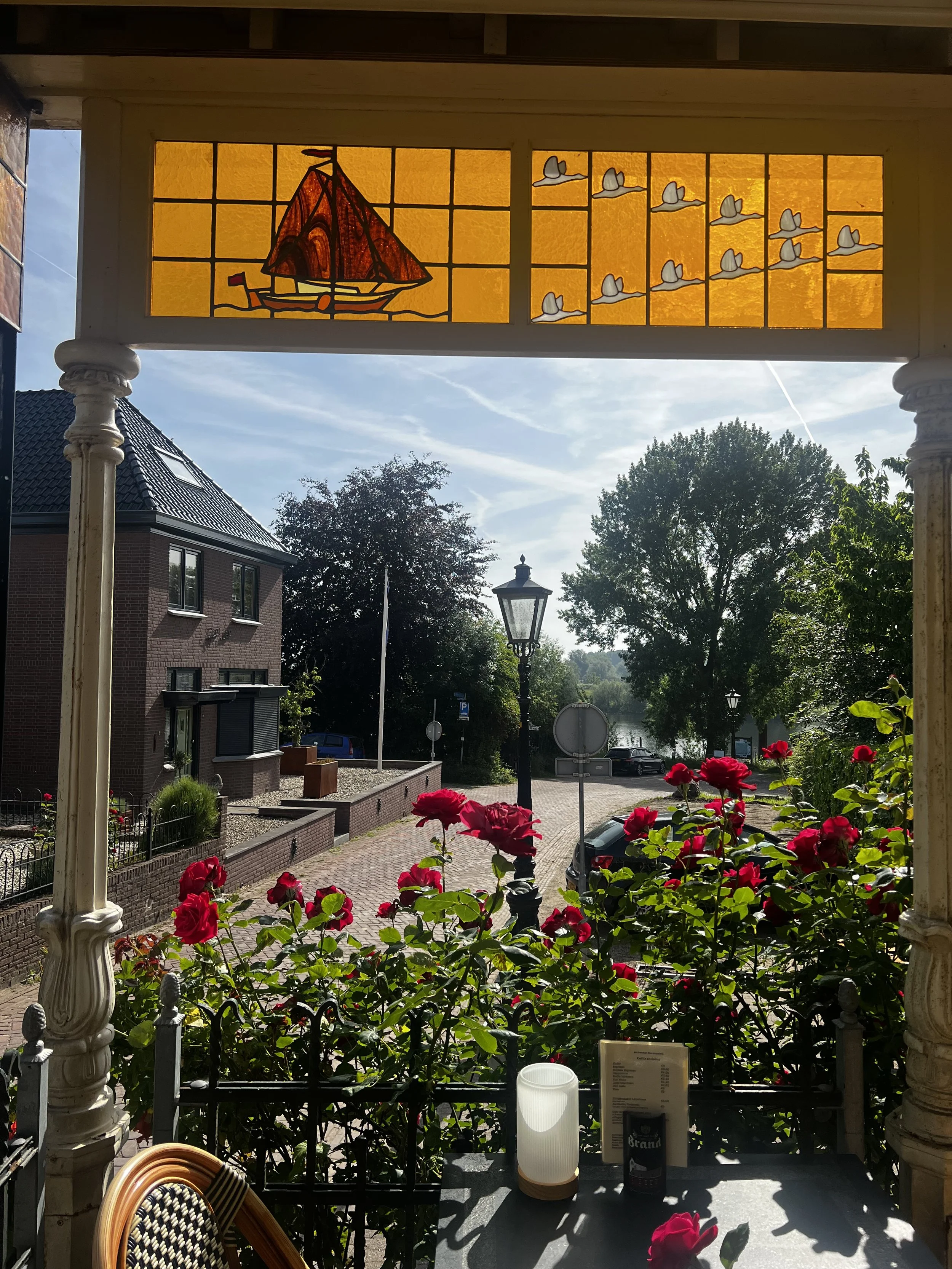 Uitzicht vanaf een terras met roze bloemen voor een raam met een glas-in-lood venster met een zeilboot en vliegende vogels, met een straat, bomen en een waterpartij op de achtergrond.