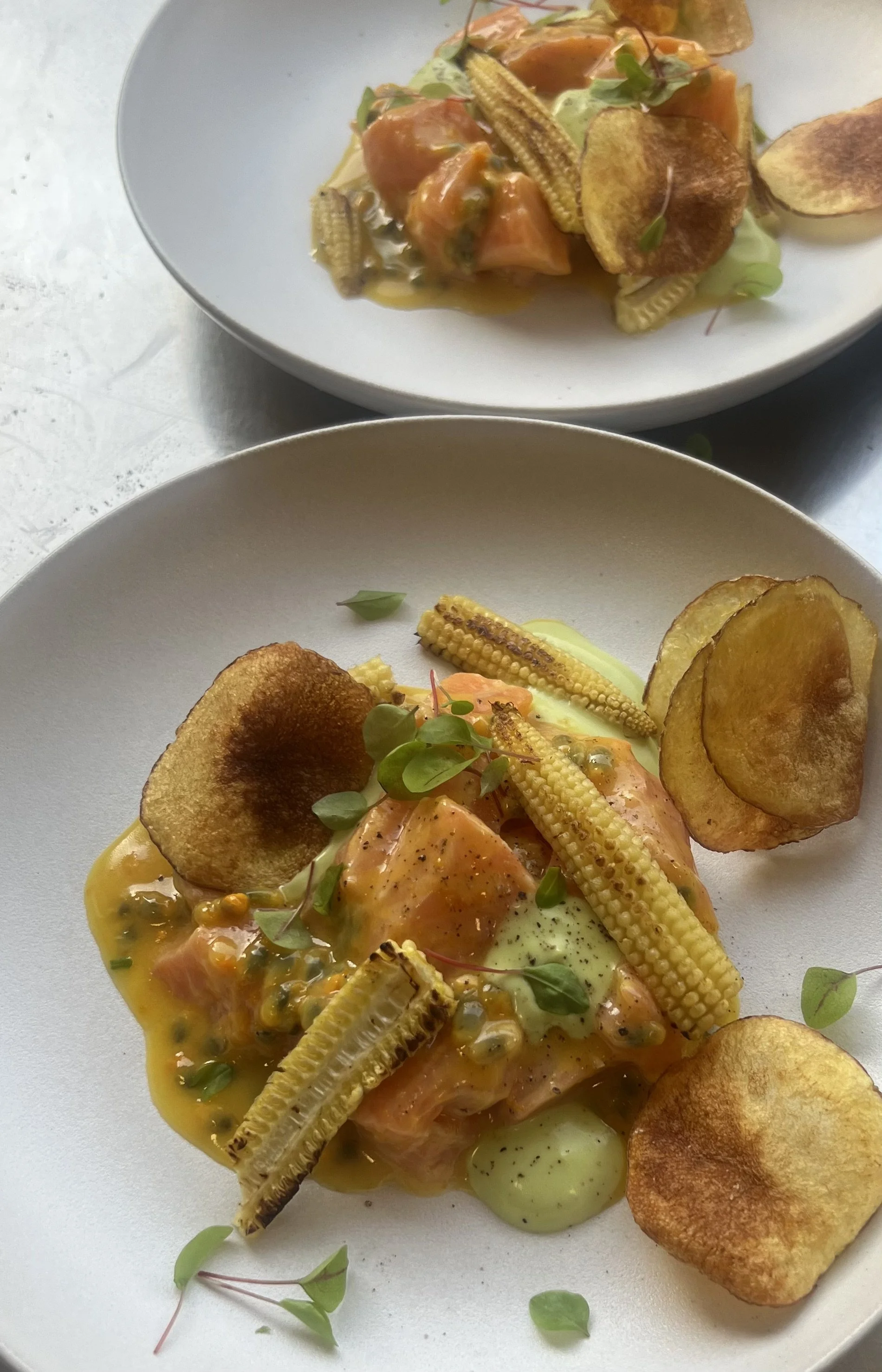 Gepromoot gerecht met stukjes gerookte zalm, gepofte aardappel, mini maïskolven, en een saus met groene kruiden, geserveerd op witte borden.