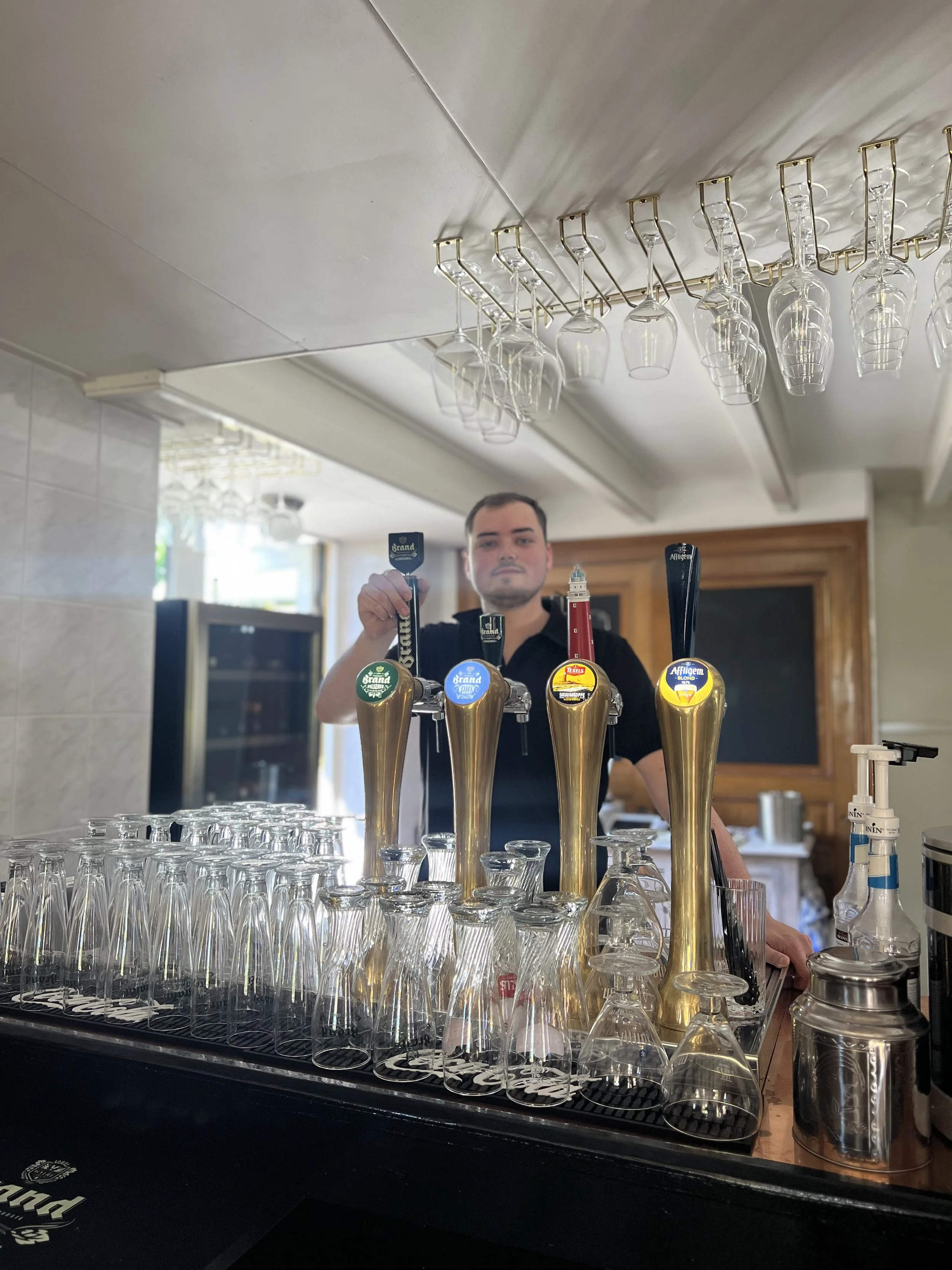 Een bar met bierkranen en glazen, een man die achter de bar staat en een ophangrek met wijnglazen aan het plafond.