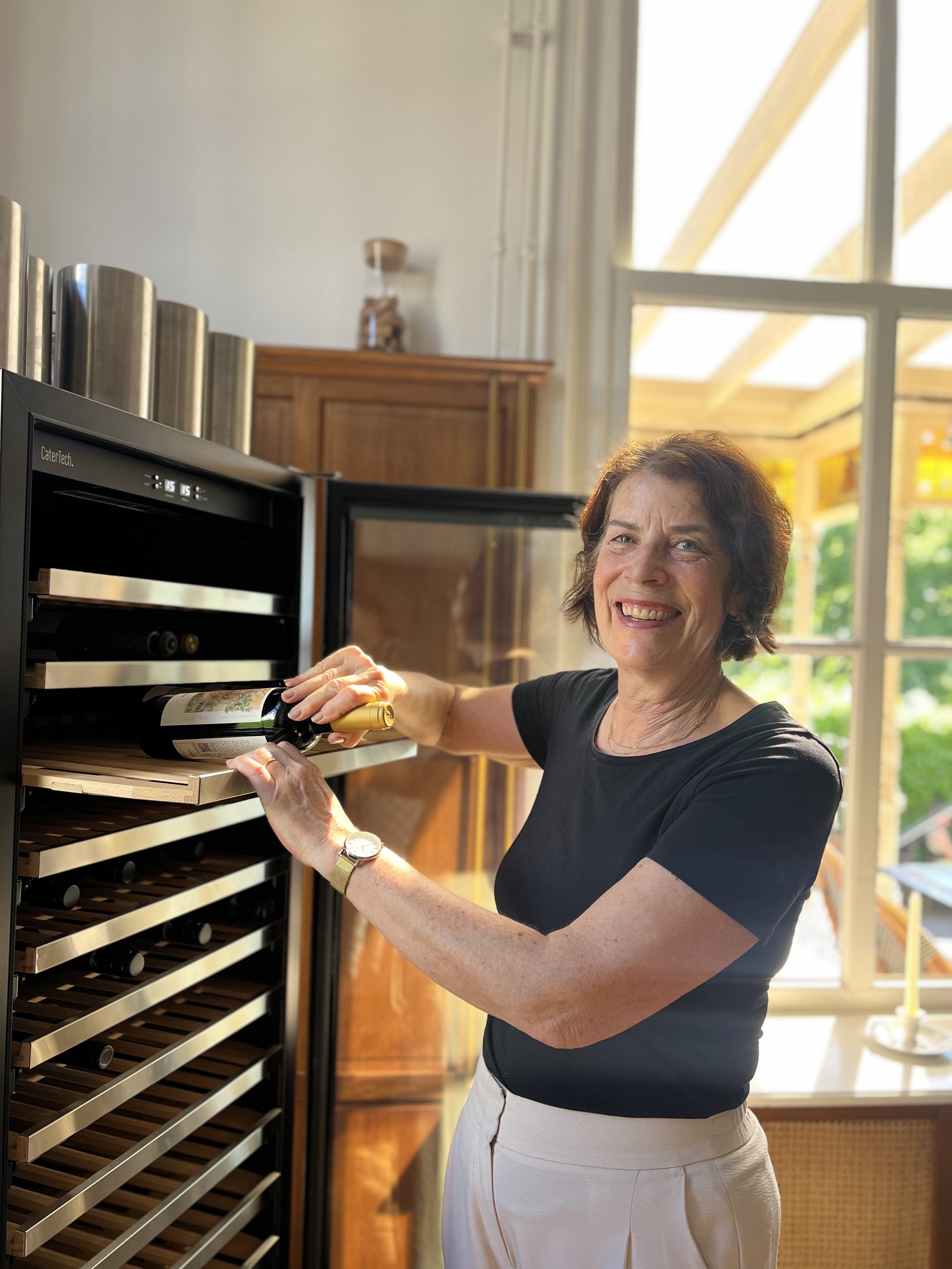 Vrouw plaatst een fles wijn in een wijnkoelkast in een keuken met grote ramen en houten vloer.