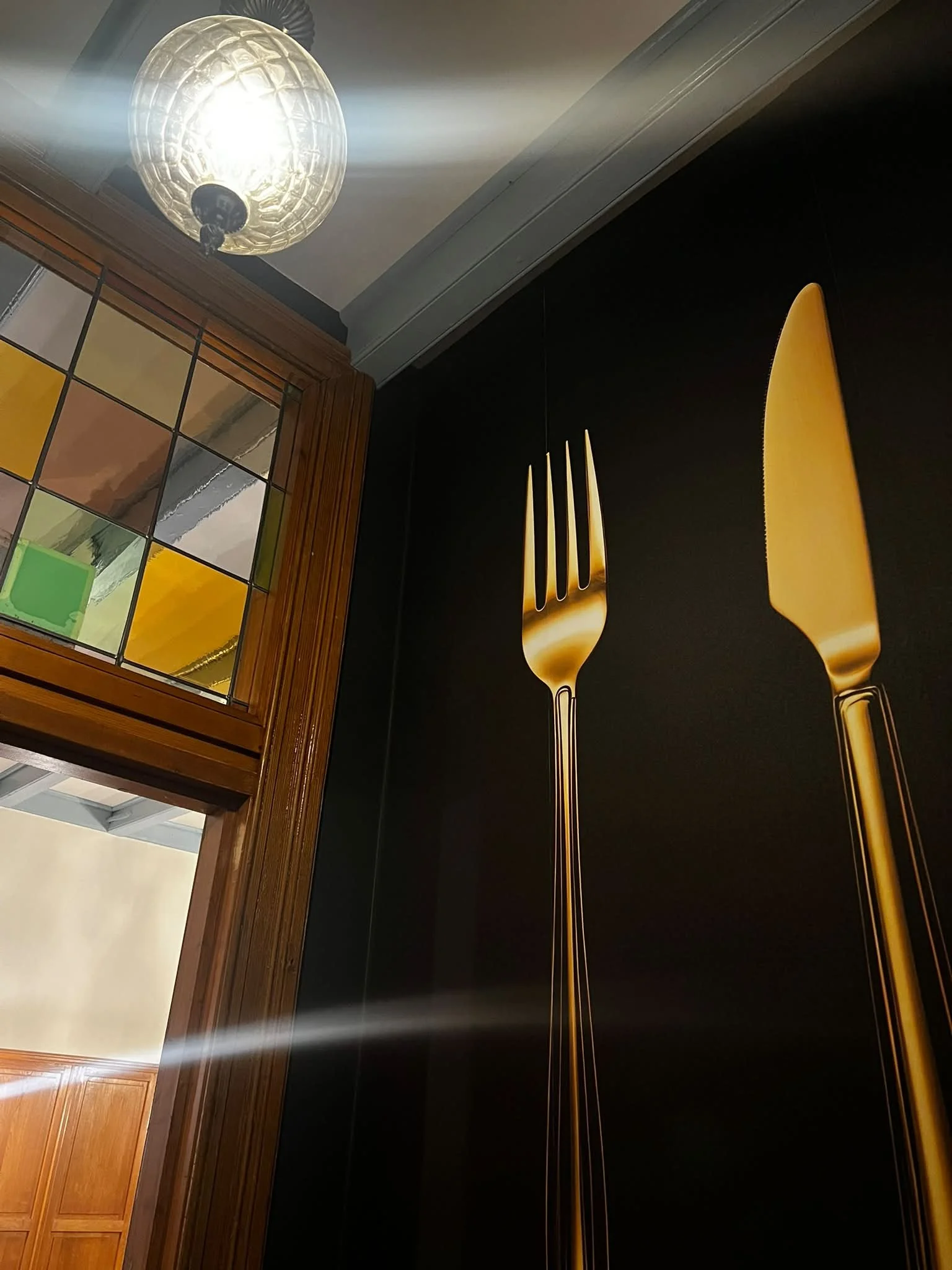 Een binnenruimte met een verlichte plafondlamp, een houten deur en een muur met een muurkunstwerk van een gouden vork en mes op een zwarte achtergrond.