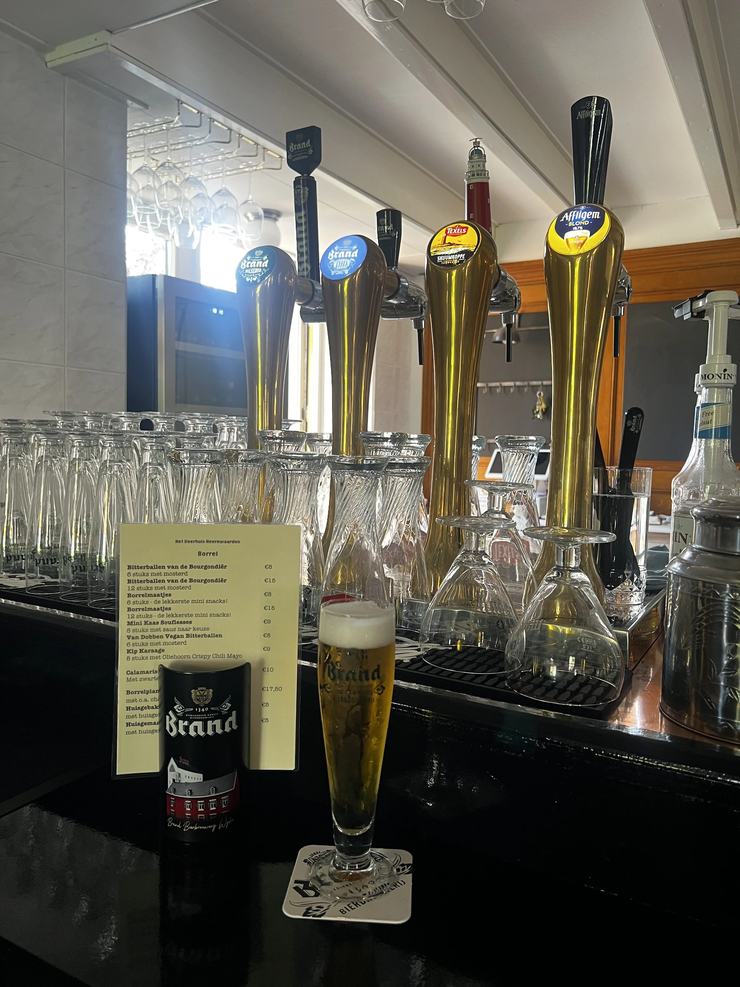 Een bar met tapkranen met verschillende soorten bier en een gevuld bierglas op de counter. Er hangt een menukaart en er staan lege glazen klaar.
