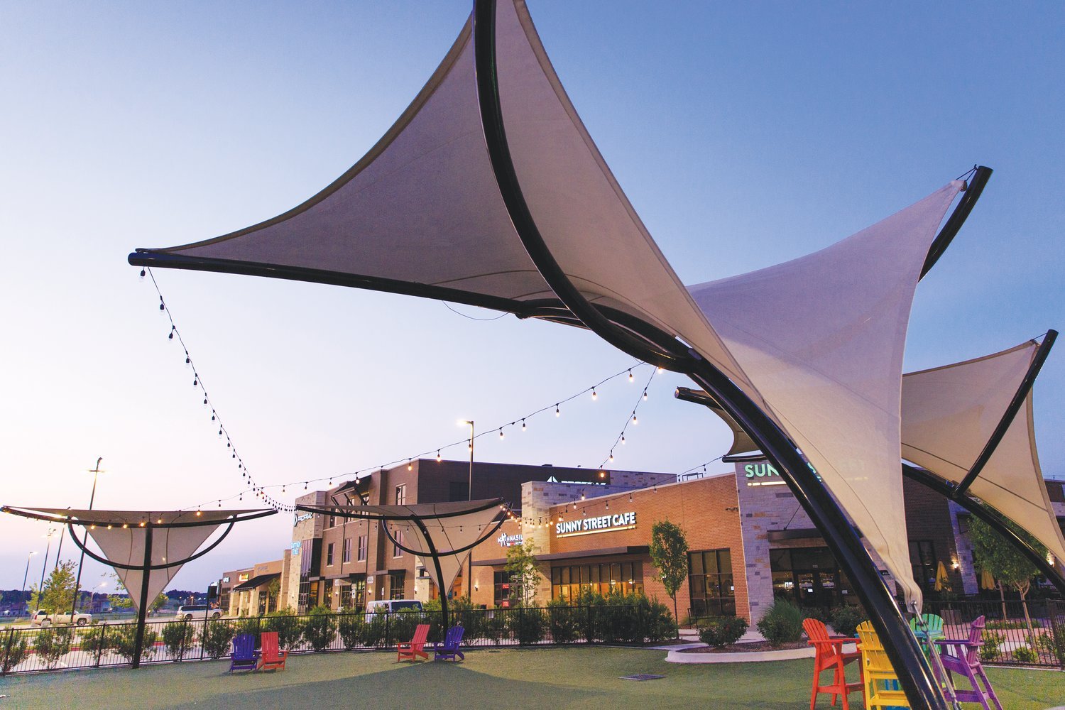 Outdoor area with colorful chairs, shade structures, string lights, and a building with a sign that reads 'Sunny Street Cafe' at dusk.