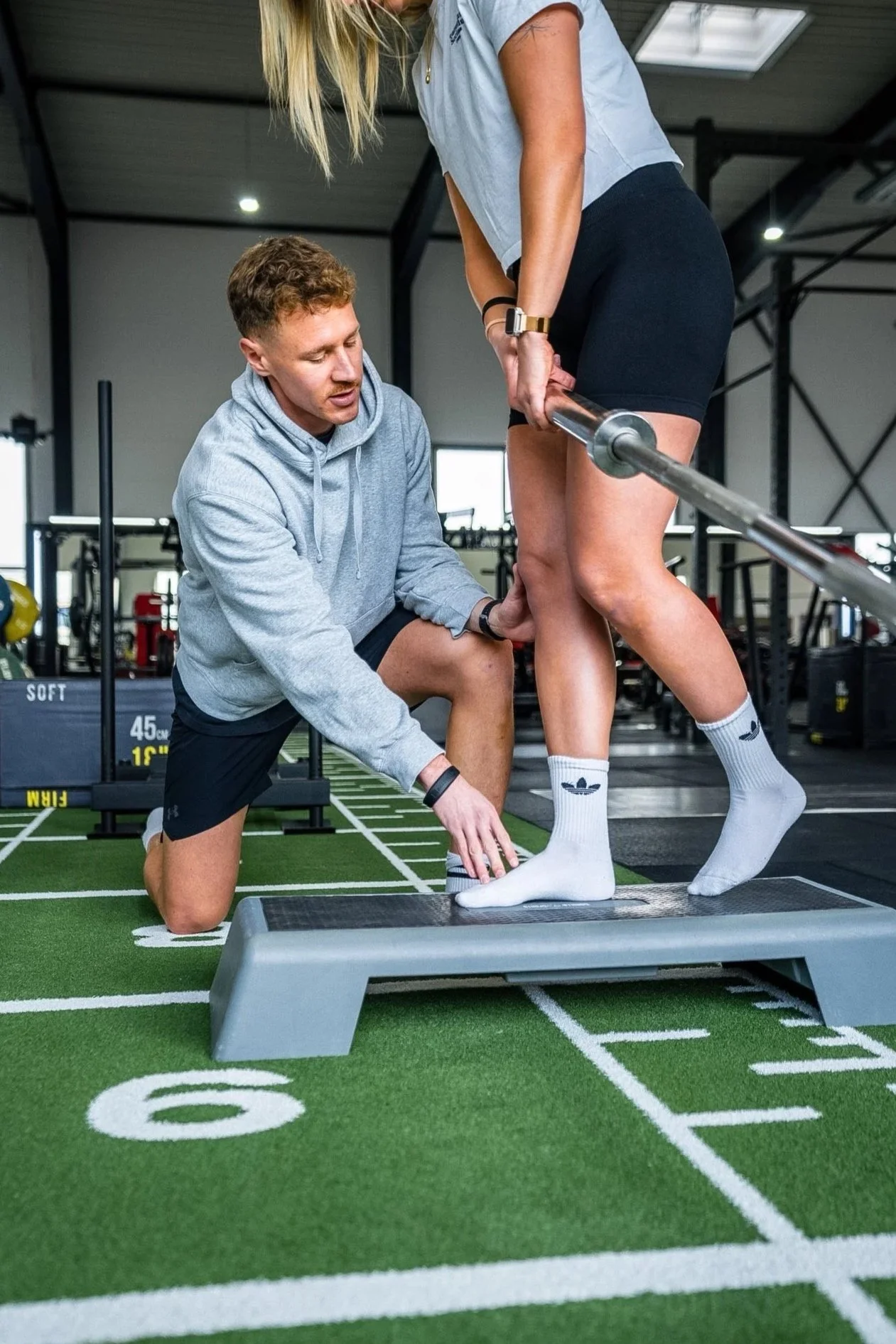 Ein Mann führt eine Übung im Fitnessstudio mit einer Frau durch. Er ist auf einem Knie, während sie auf einer Plattform steht, mit einer Langhantel auf ihren Oberschenkeln.