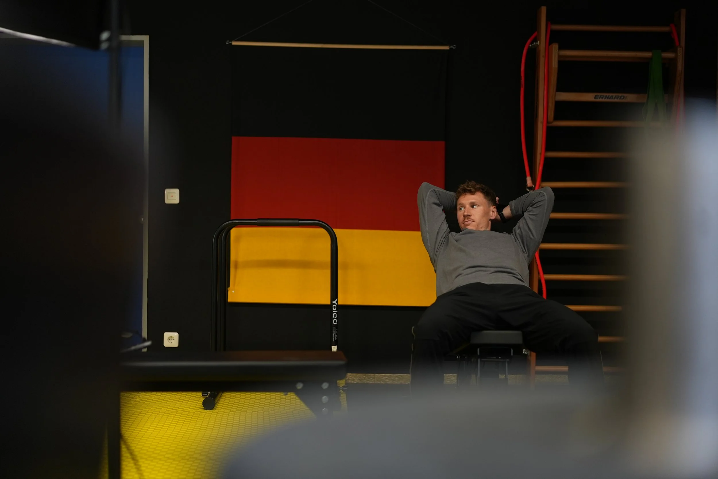 Ein Mann liegt auf dem Boden in einem Fitnessraum, entspannt mit den Händen hinter dem Kopf, mit einer schwarzen Wand im Hintergrund, einer roten und gelben Wanddekoration, einer Holzleiter an der Wand, und Trainingszubehör wie einem Vertébar-Trainingsgerät.