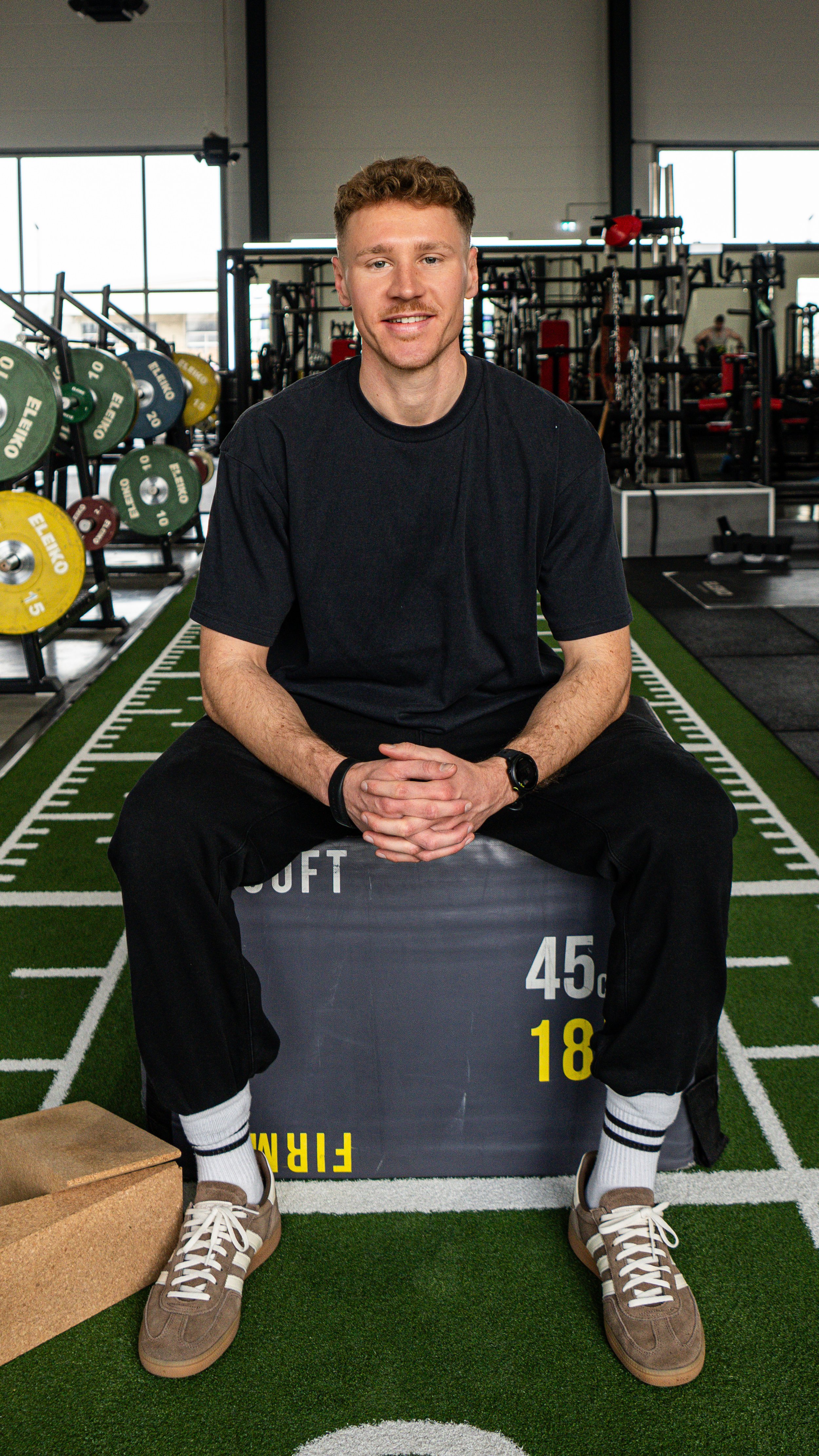 Ein Mann sitzt auf einem Plyo-Box im Fitnessstudio, umgeben von Gewichten, Trainingsgeräten und Fitnesszubehör, trägt schwarze Kleidung, Sportschuhe und schaut in die Kamera.