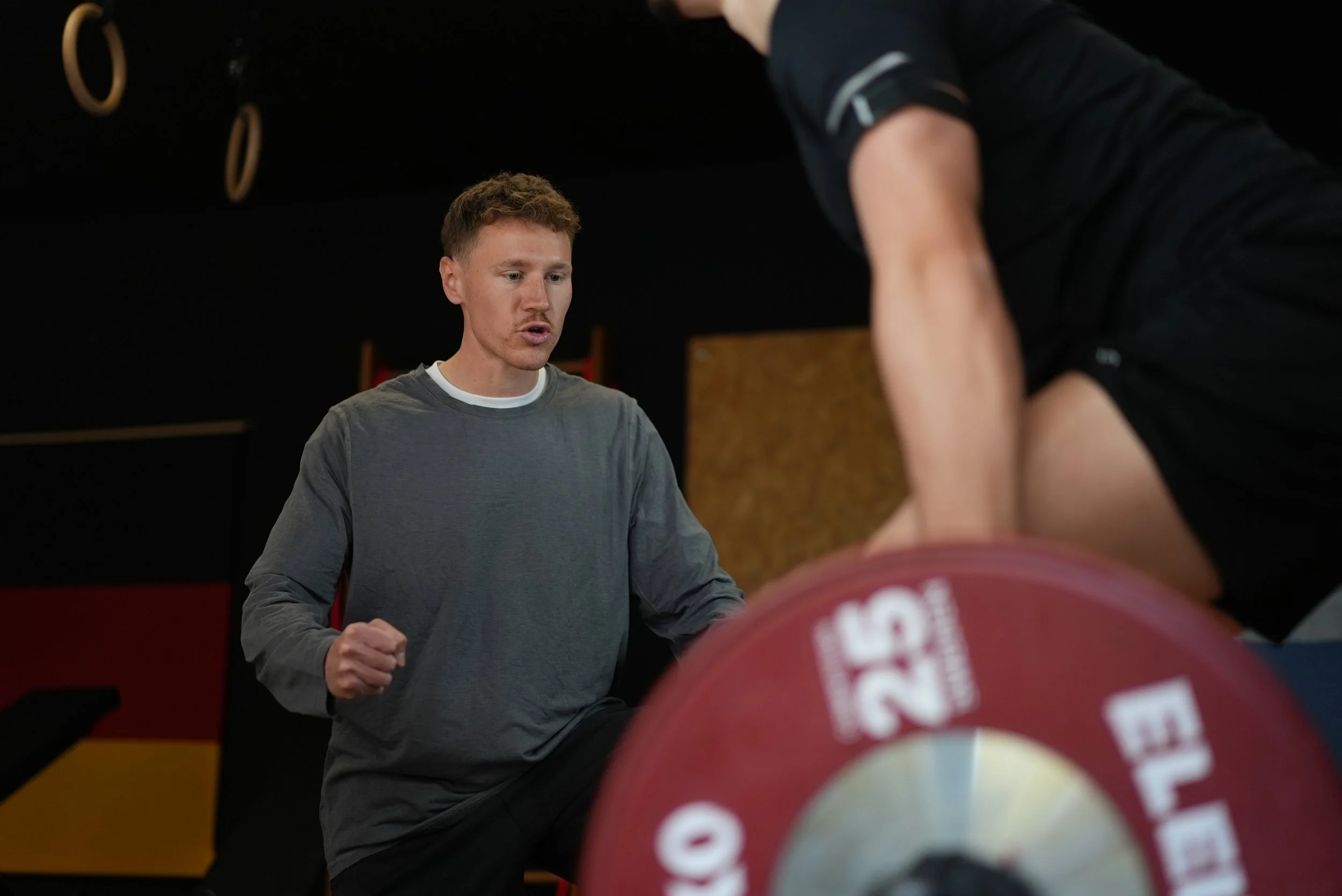 Zwei junge Männer im Fitnessstudio, einer hebt eine Hantel, der andere beobachtet.