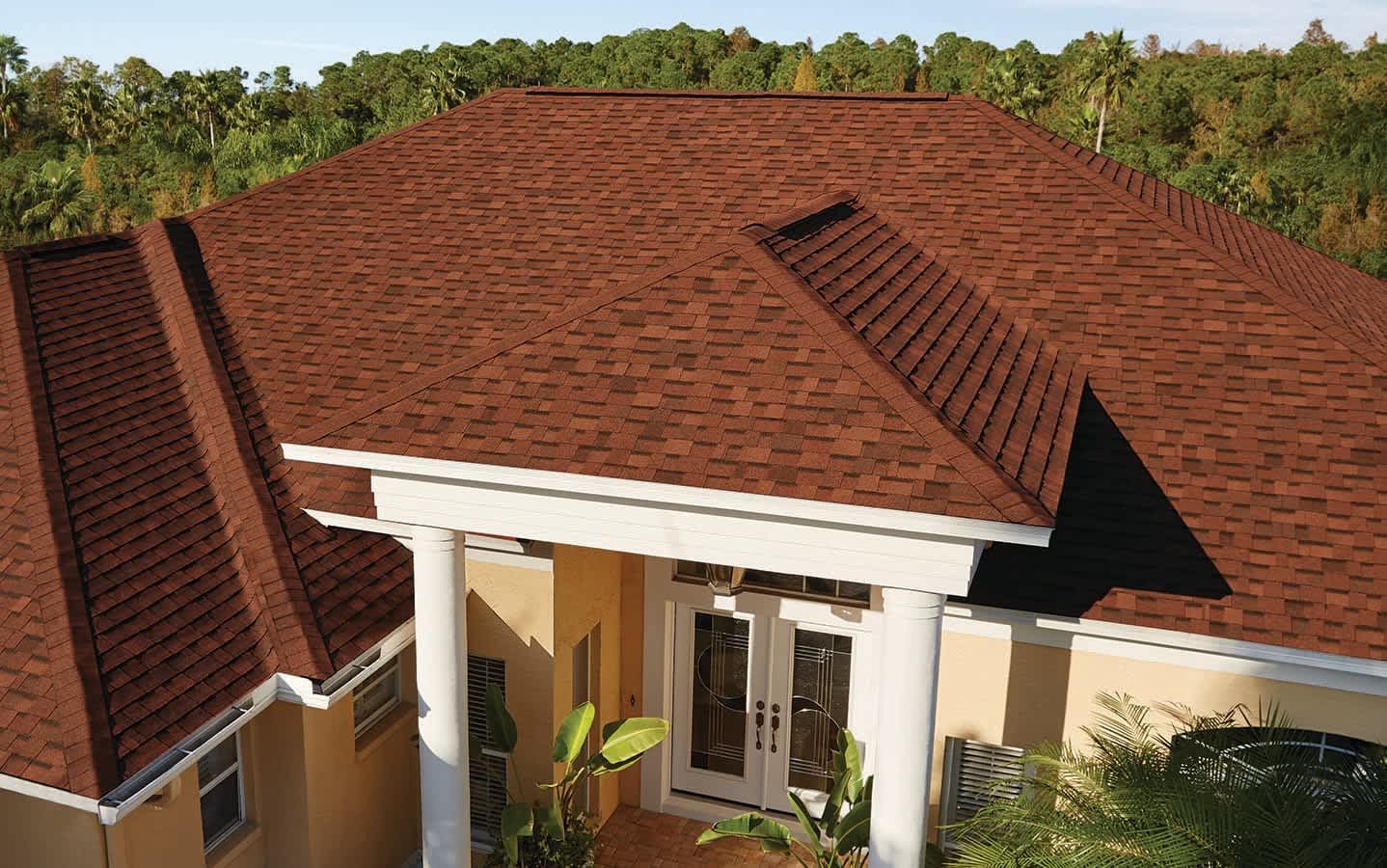 A house with a red shingle roof, white columns at the entrance, yellow exterior walls, and tropical plants near the door, set against a backdrop of green trees.