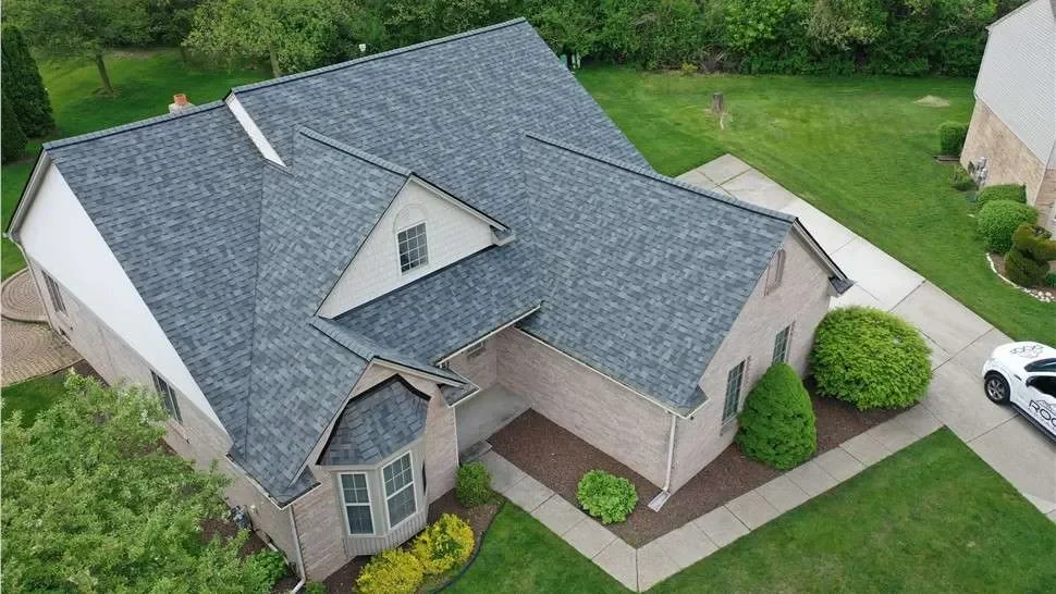 A house with a multi-gabled roof, beige brick exterior, and white trim, surrounded by a well-maintained lawn and shrubbery, with a driveway and a white vehicle parked on it.