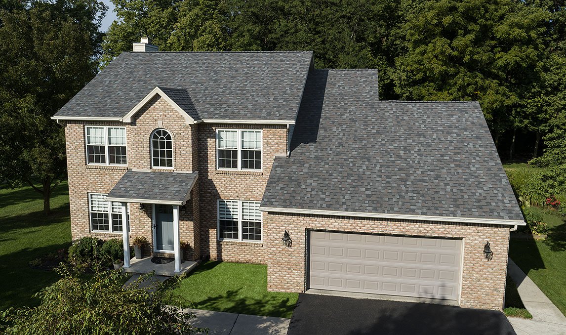 A two-story brick house with a gray shingle roof, front porch with white columns, and a two-car garage. Surrounded by green lawn and trees.