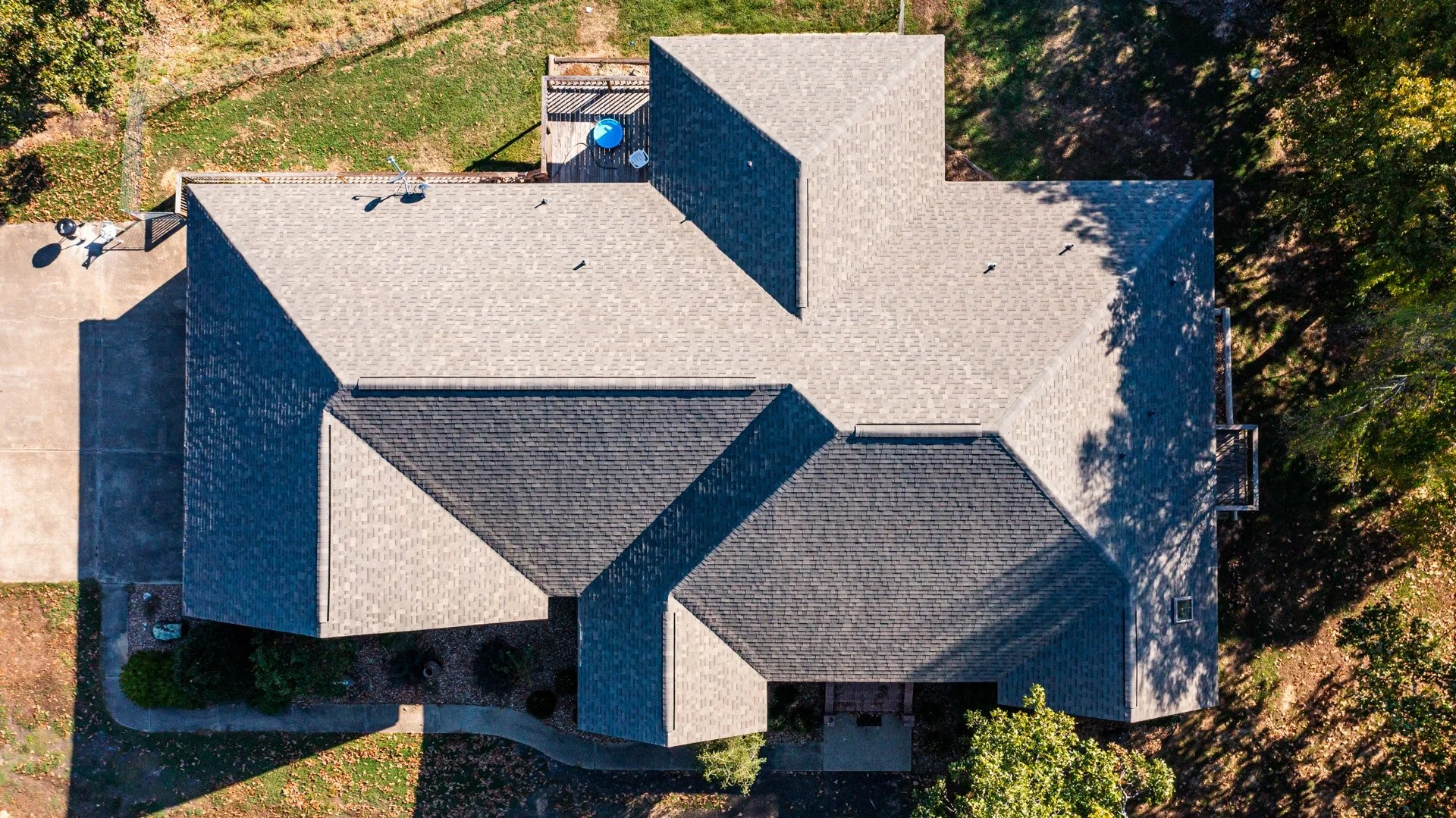 Aerial view of a house's roof with multiple gables, surrounded by trees and a backyard with a deck, patio, and lawn.