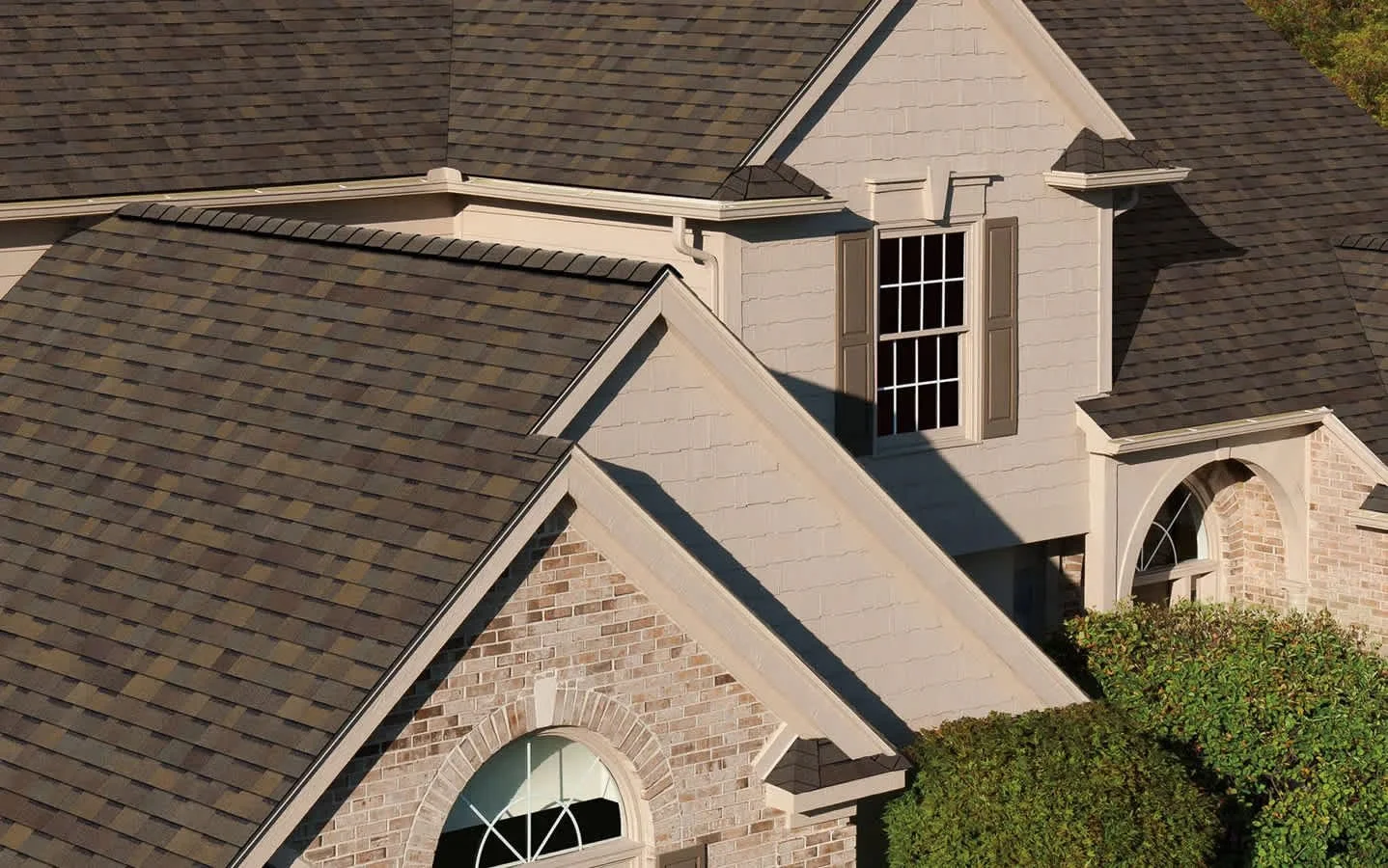 Close-up of a residential house's roof and upper windows, with brown shingles and beige siding.