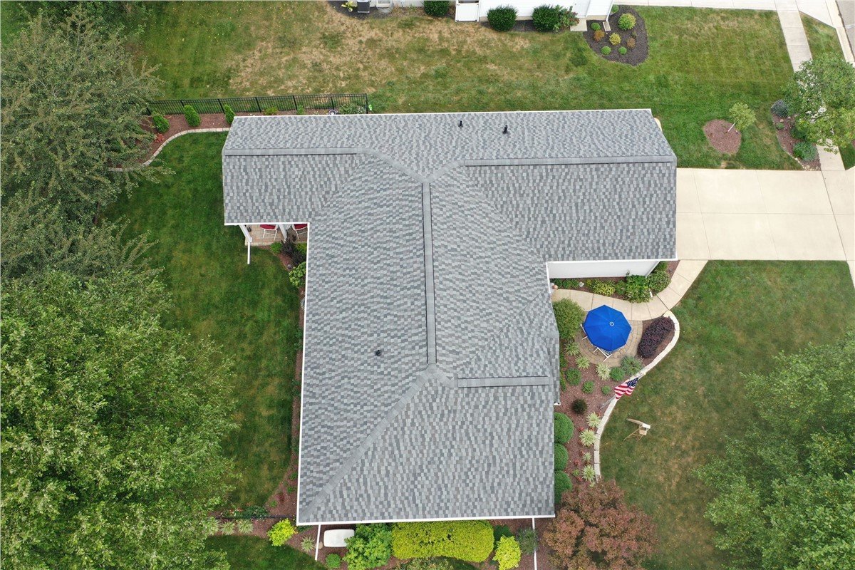 Aerial view of a house with a gray shingled roof, a well-maintained backyard with green grass, trees, a side patio with a blue umbrella, and a concrete driveway.