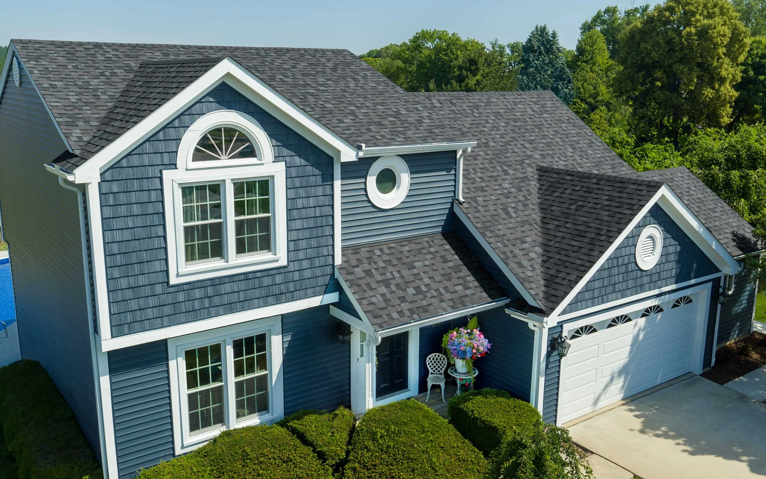 A blue two-story house with white trim and a gray shingled roof, featuring large windows, circular window accents, and a small porch with a white table, chairs, and flower arrangement.