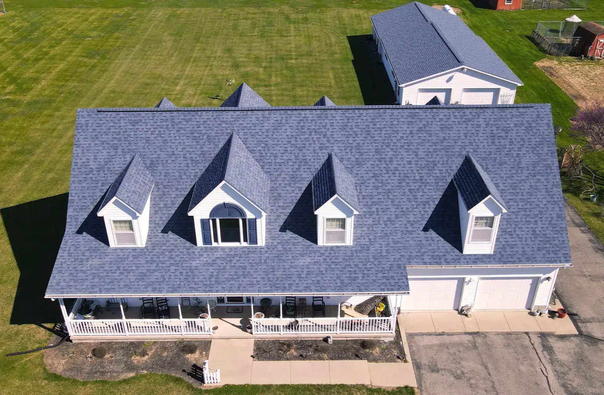 Aerial view of a white house with a gray shingle roof, dormer windows, and a large front porch with outdoor furniture, surrounded by a well-maintained green lawn and additional structures, including a detached garage and a garden shed.