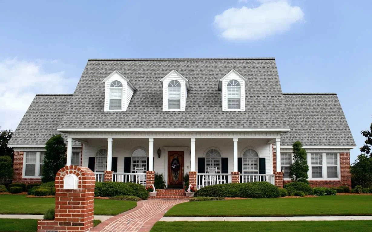 Front view of a large, two-story house with a gray shingle roof, white wooden siding, black shutters, and a brick foundation. The house has a front porch with white columns, a wooden front door, and a brick walkway leading to the sidewalk. The front 