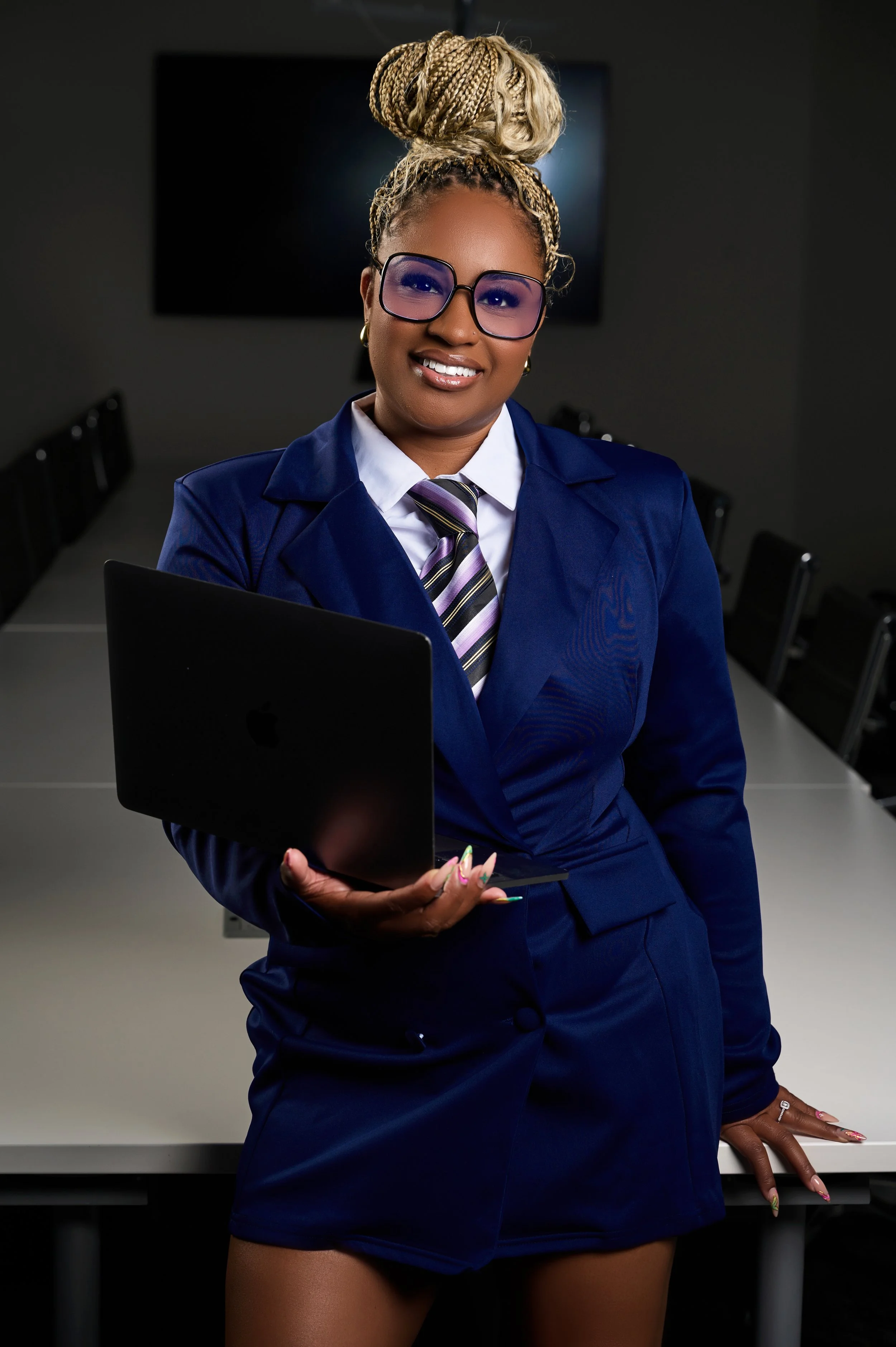 A woman dressed in a blue business suit and tie, wearing large purple sunglasses, holding a closed laptop, standing in a conference room with a large blank screen behind her.