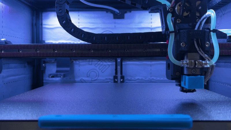 A 3D printer printing on a flat build platform, with mechanical parts and tubing visible.