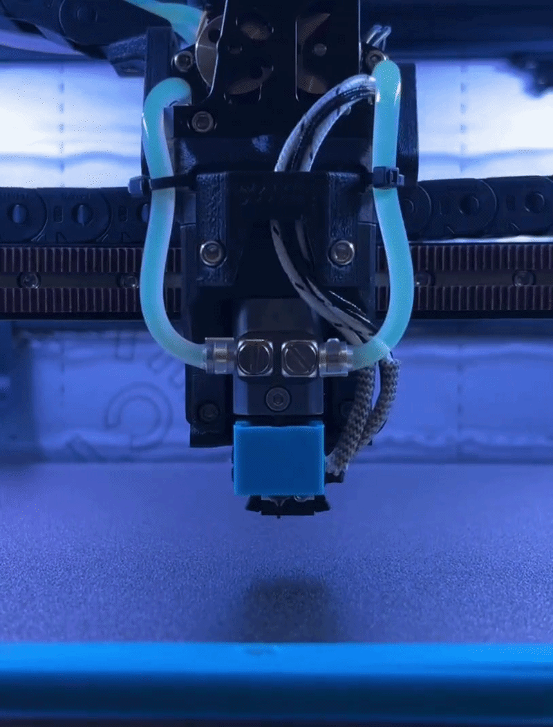 Close-up of a 3D printer nozzle in operation, with blue plastic filament being extruded, under blue lighting.