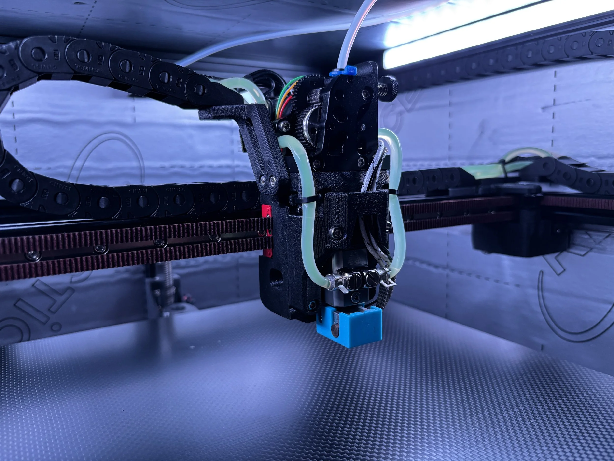 Close-up of a mechanical 3D printer's moving part with black plastic chain links, green tubing, multicolored wires, and metal gears, set against a metallic background.
