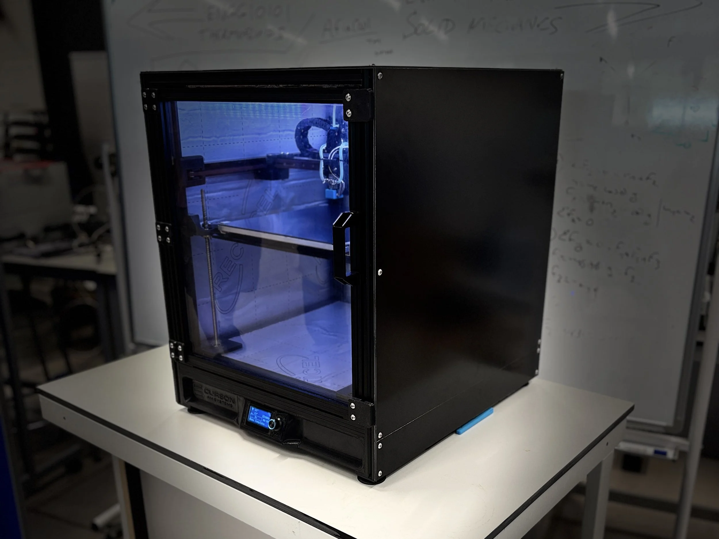 A black 3D printer with a transparent front door, placed on a white desk in a laboratory or workshop setting.