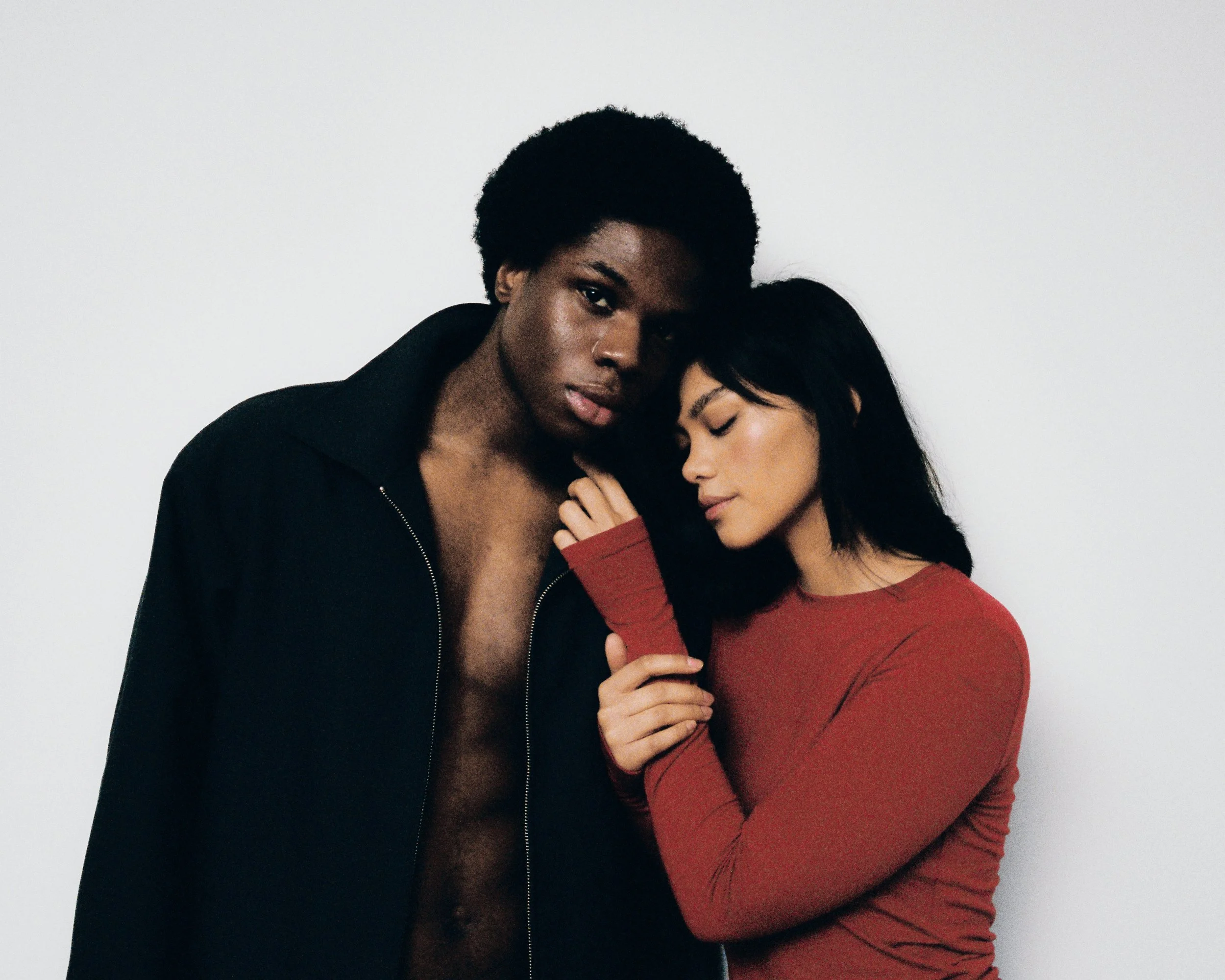 Two women, one with dark skin and curly short hair, wearing a black jacket, and the other with light skin and long dark hair, wearing a red top, are close together with their eyes closed, embracing each other softly.