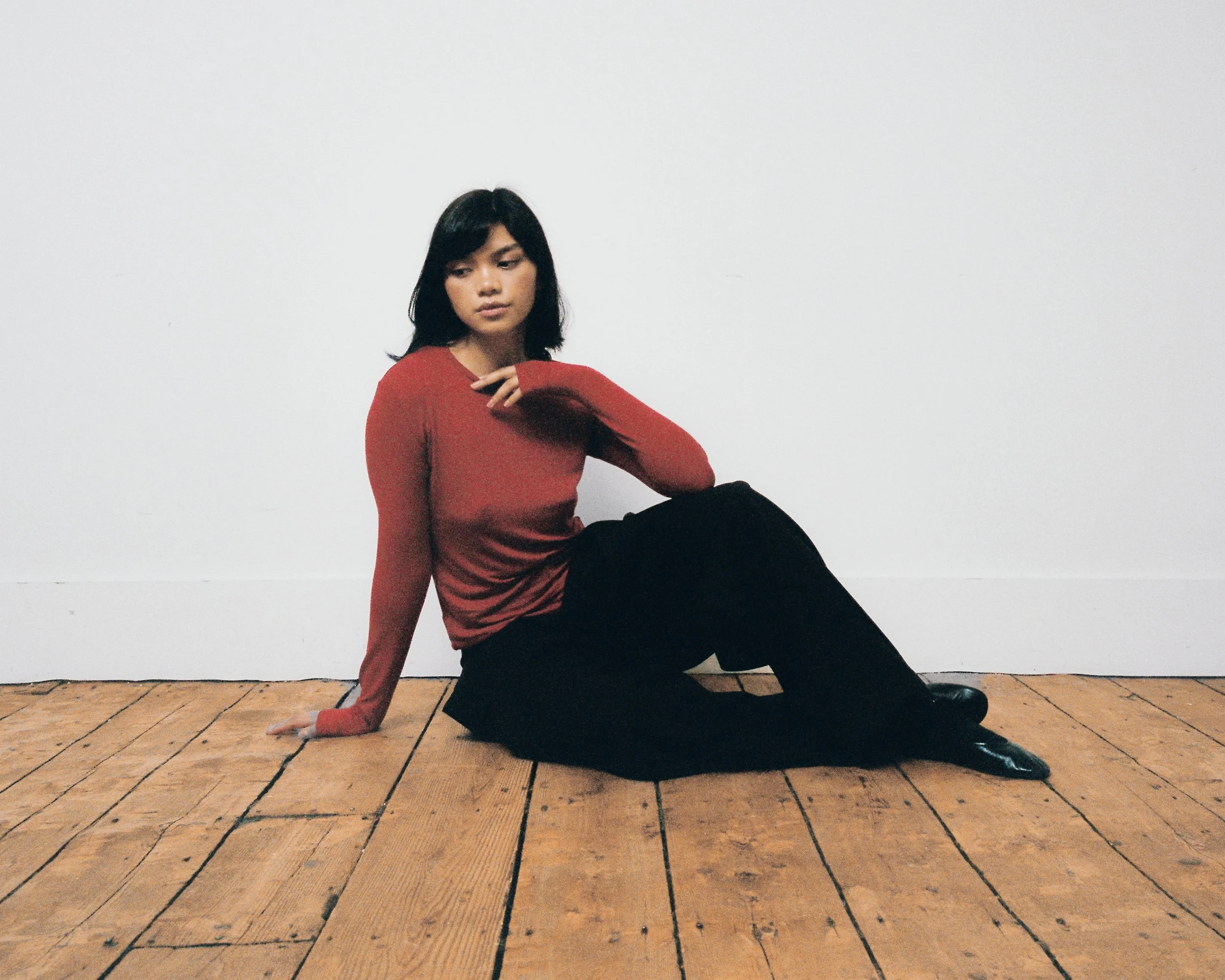 A woman with dark hair wearing a red long-sleeve shirt and black pants sitting on a wooden floor against a plain white wall, looking downward with her left hand on her lap and her right hand near her neck.