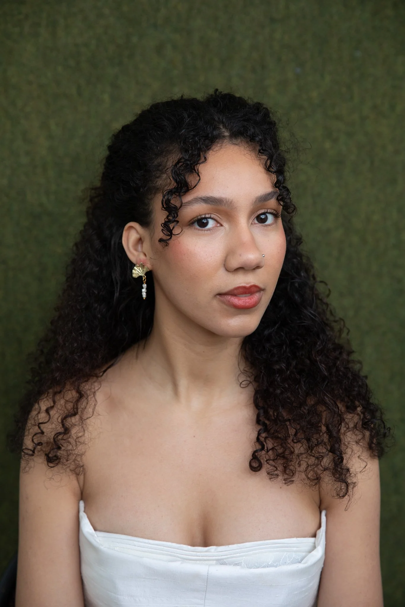 Close-up of a young woman with curly dark hair, wearing a white dress and gold earrings, against a green textured background.