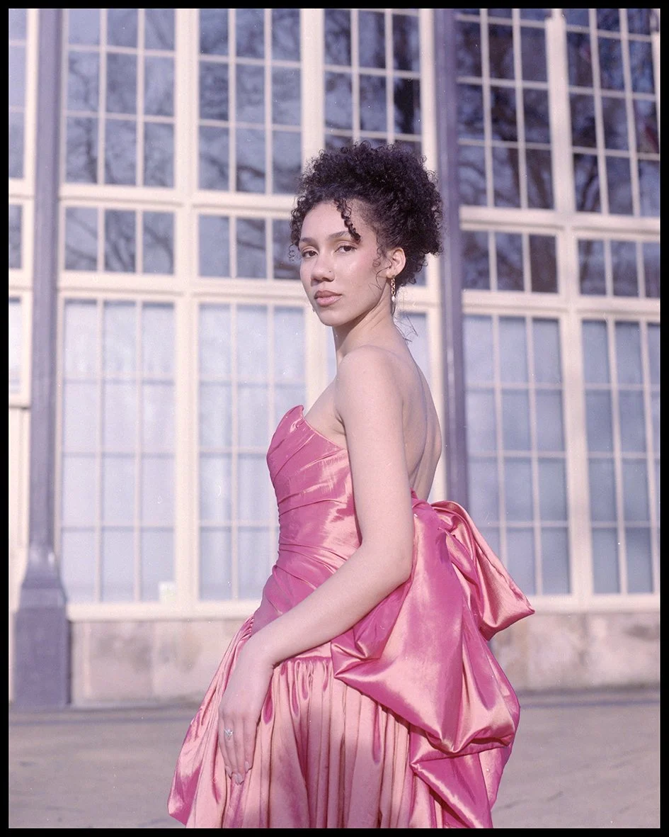 A young woman in a pink satin gown standing in front of a large glass window.