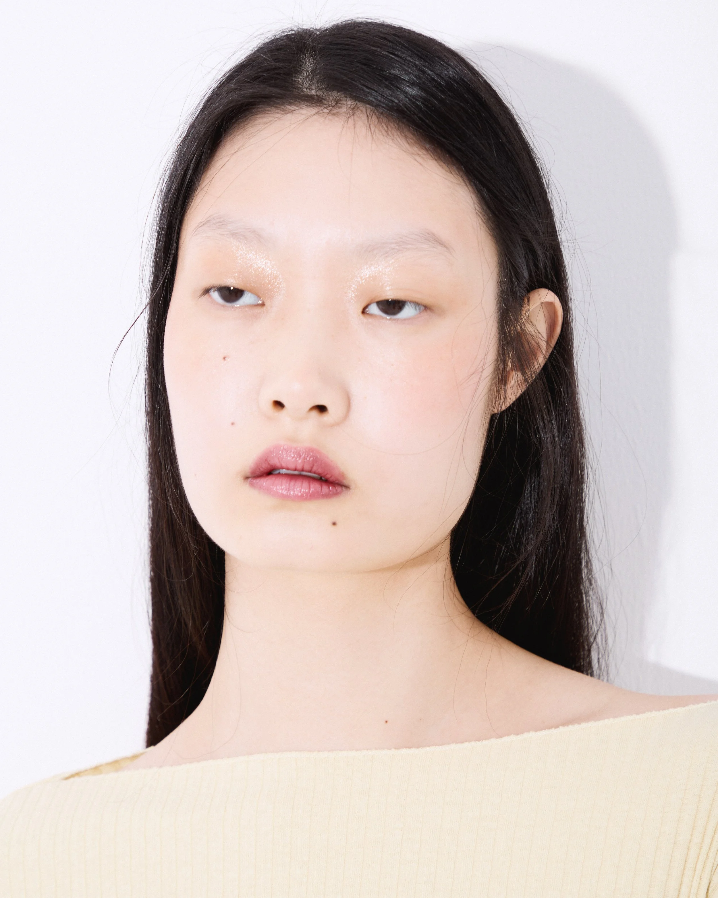 Close-up of a woman's face with dark hair, minimal makeup, and a neutral expression against a plain white background.
