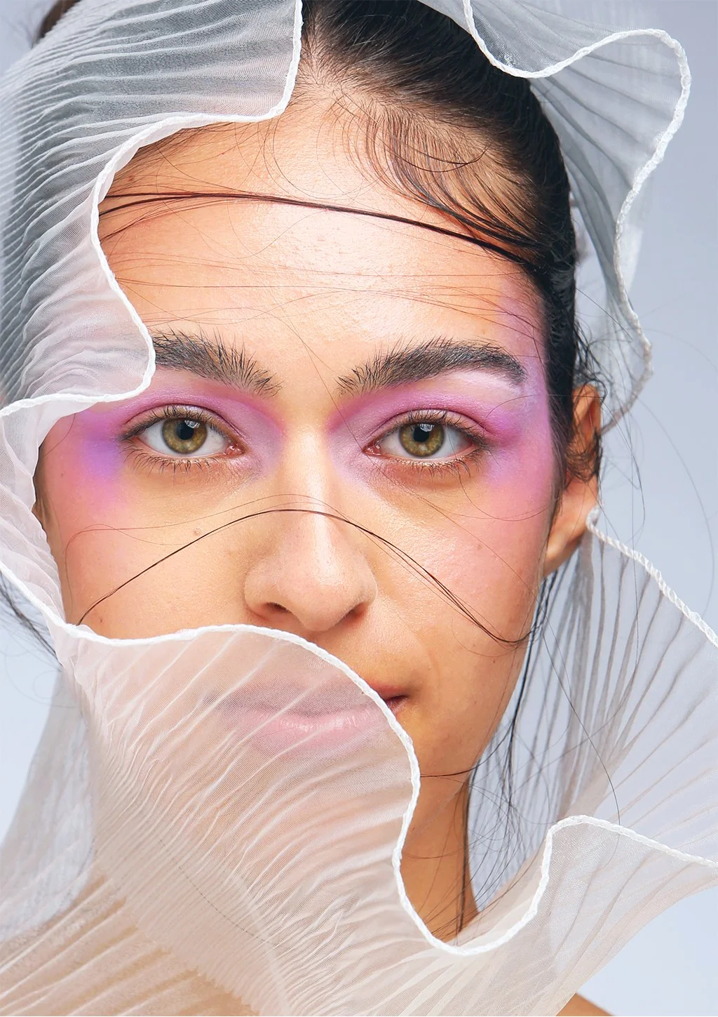 A woman with light brown eyes and pink eyeshadow, partially covered by a sheer, pleated fabric with a ruffled edge, framing her face.