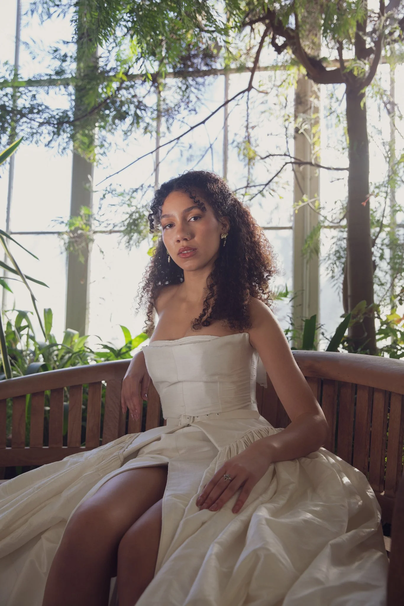 A woman with curly hair and earrings sitting on a wooden bench in a sunlit room with tall windows and lush green plants.