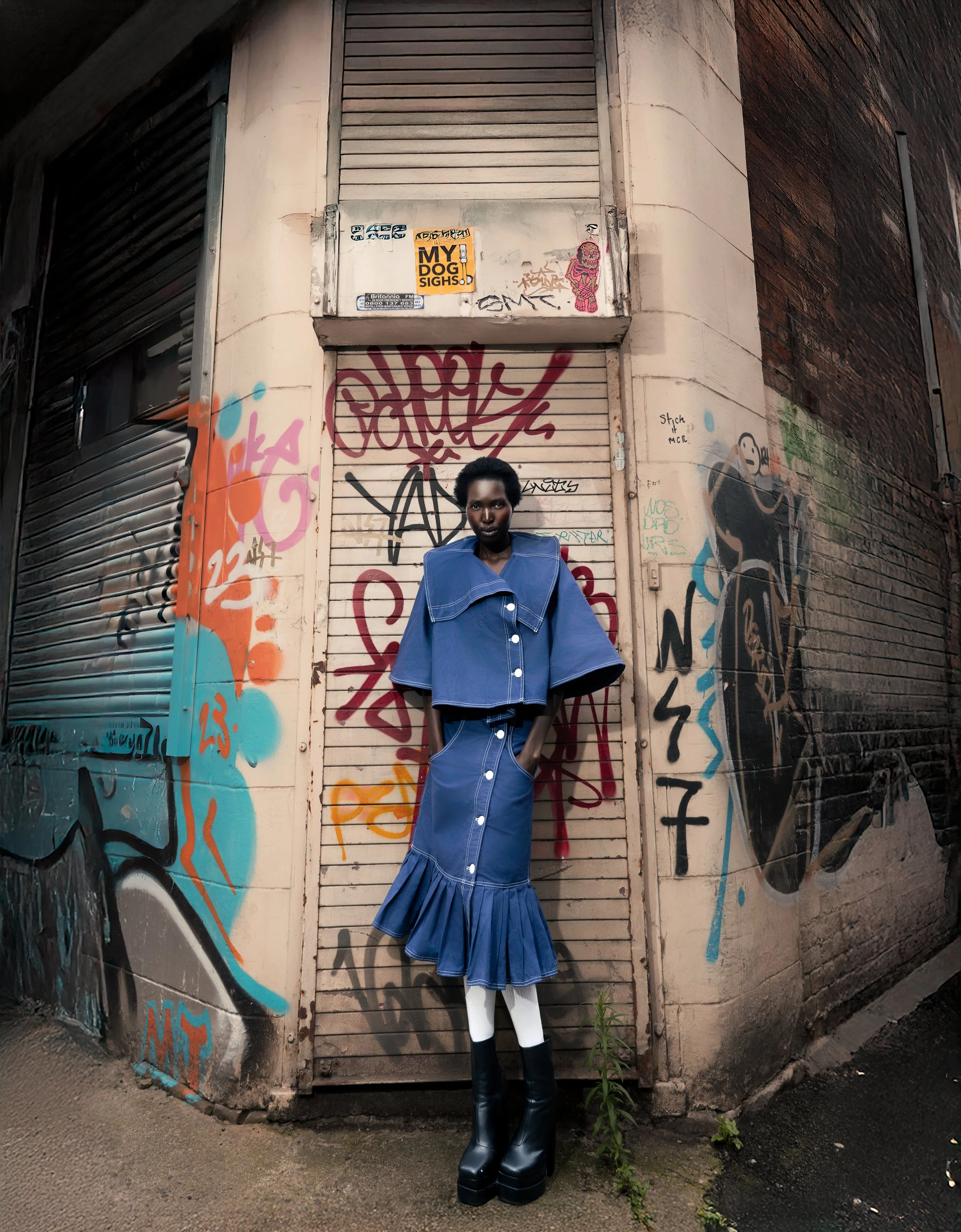 A person with dark skin and short curly hair facing forward stands in front of a graffiti-covered metal garage door. They are wearing a blue oversized button-up dress with a pleated skirt, white tights, and black platform boots. The surrounding walls are painted with various graffiti and stickers.