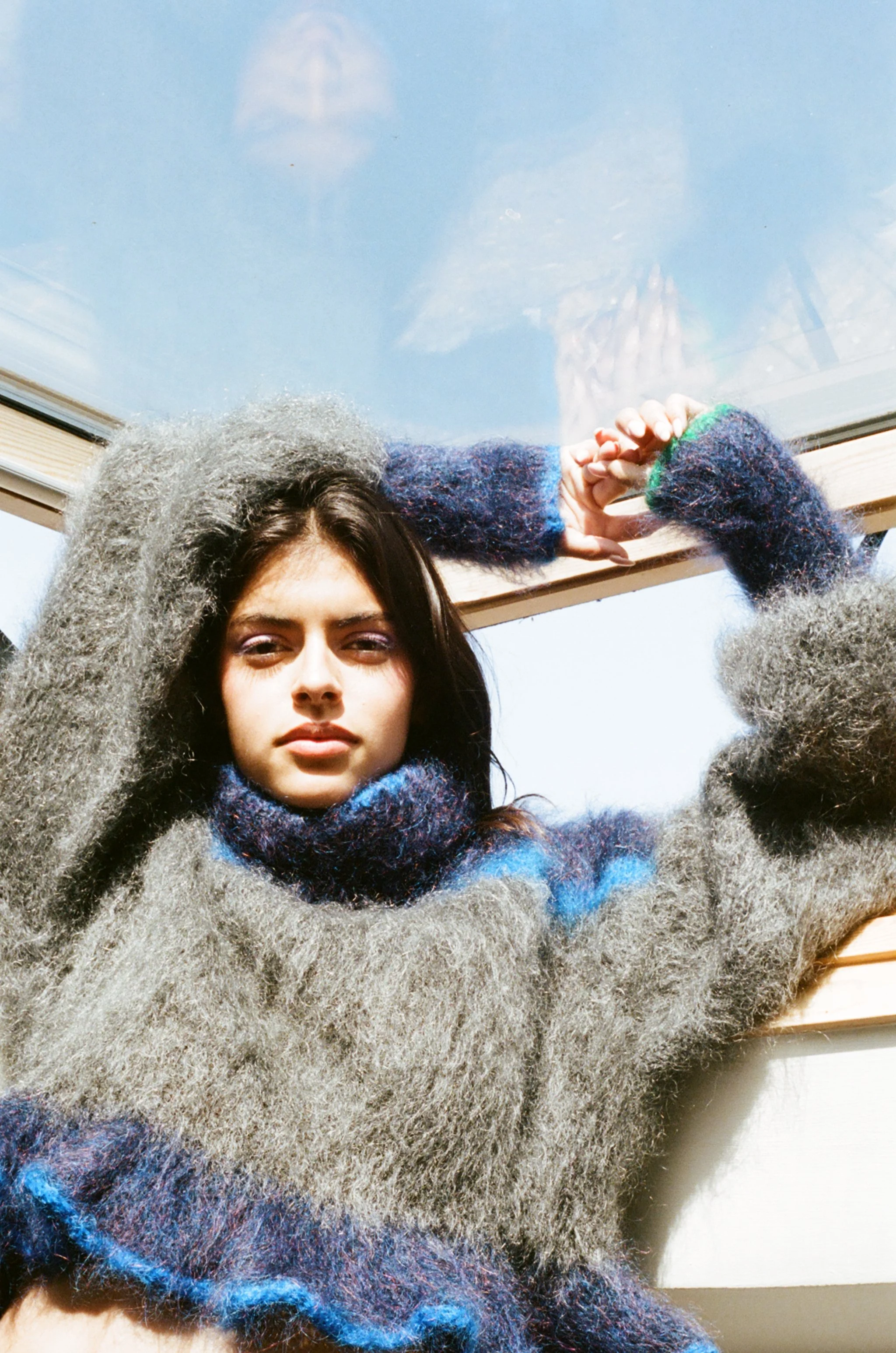 A young woman with dark hair wearing a colorful scarf, fluffy gray sweater, and fingerless mittens, is standing outdoors under a blue sky with her hands against a glass surface, looking at the camera.