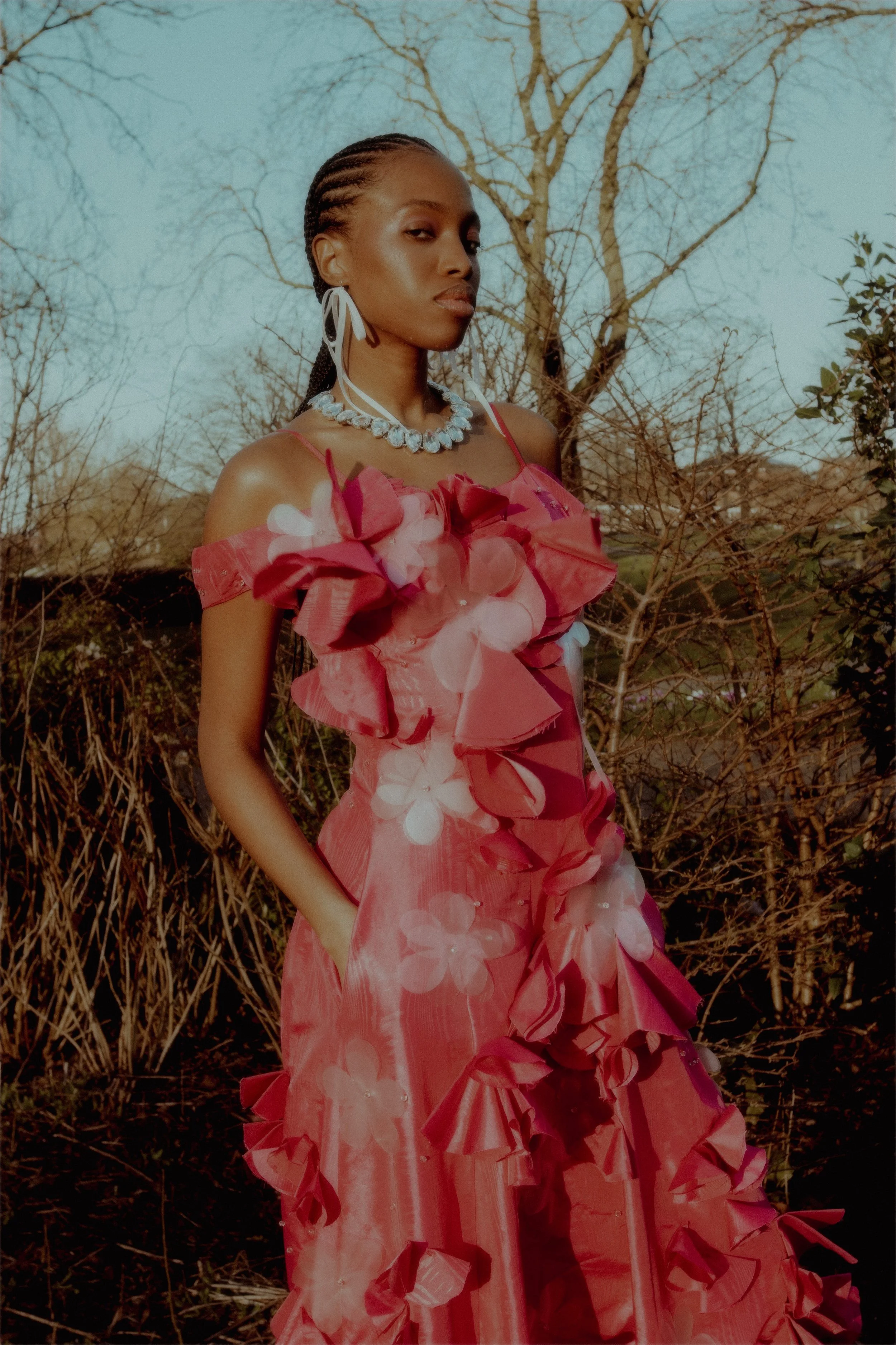 Woman in a pink floral gown with ruffles, standing outdoors with bare trees in the background.